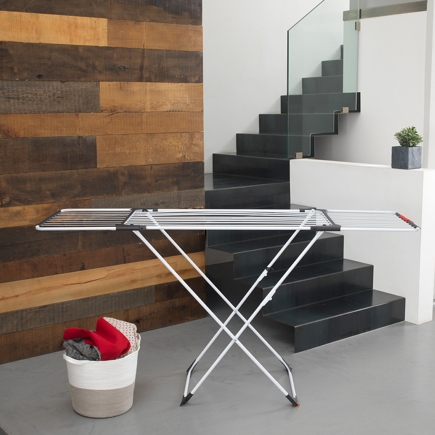 Uno stendibiancheria è di fronte a un muro di legno e a delle scale. Un cesto di vestiti è sotto. Lo sfondo è uno spazio moderno.