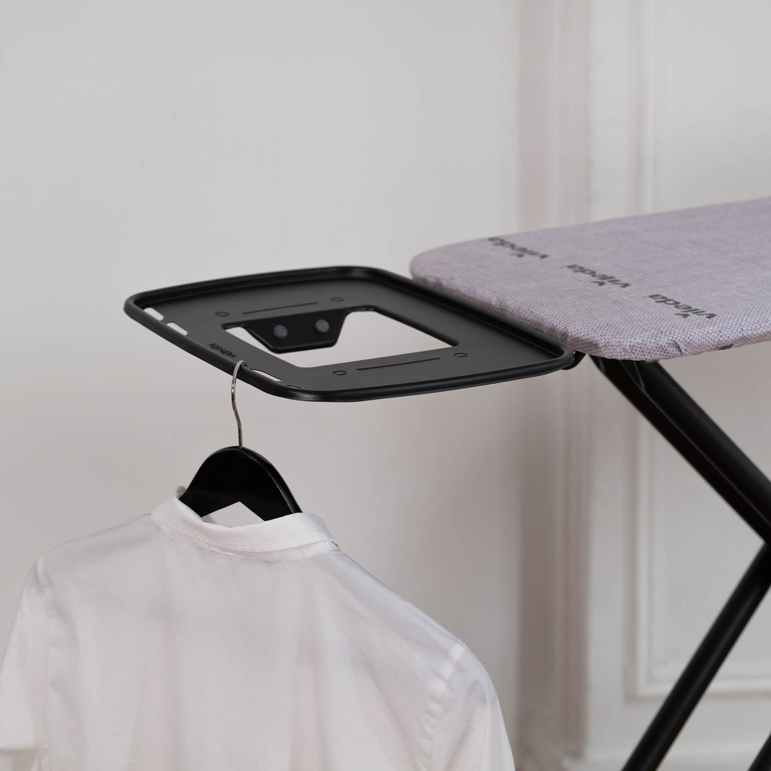 Un asse da stiro con una camicia bianca su una gruccia. L'asse è nero e grigio e il muro è bianco.