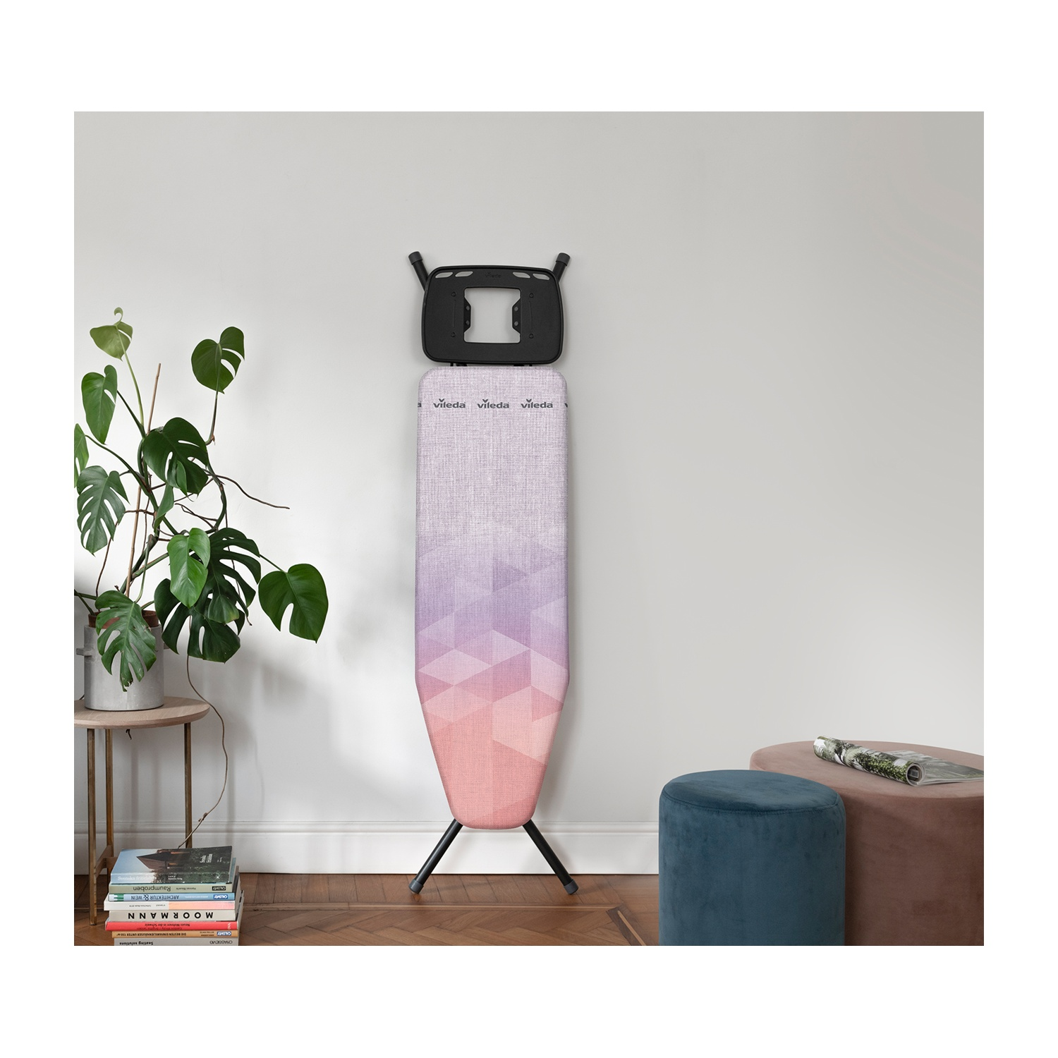 Asse da stiro con design geometrico rosa e viola, gambe nere e supporto a parete. Presenti anche pianta, sgabelli e libri.
