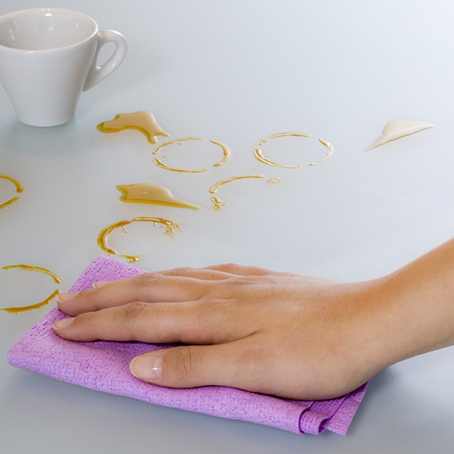 Una mano pulisce un liquido versato su un tavolo con un panno viola. Una tazza bianca è vicina. Macchie marroni indicano la fuoriuscita.