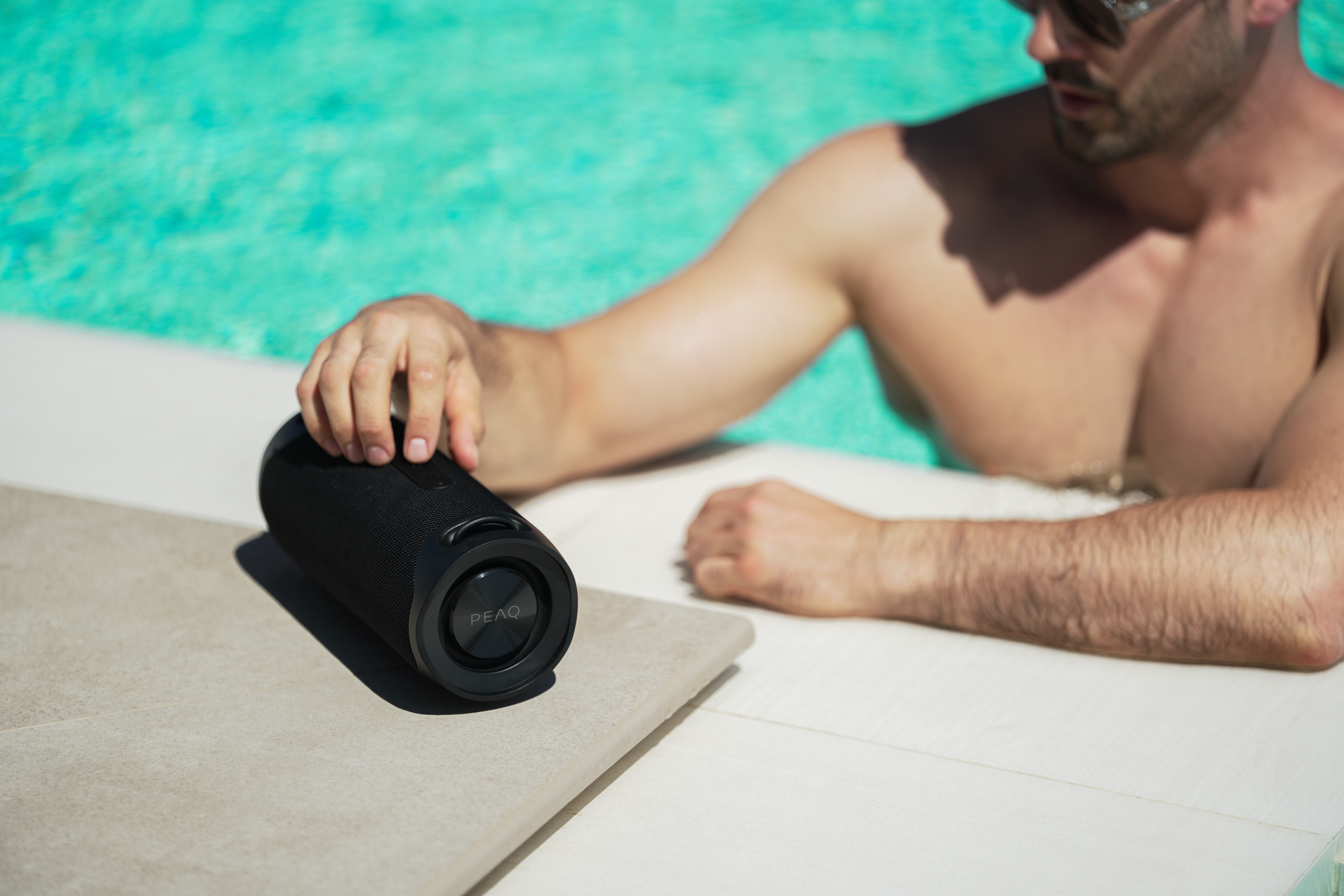 Ein Mann am Pool hält einen schwarzen Lautsprecher. Er trägt eine Sonnenbrille. Türkisfarbenes Wasser im Hintergrund.