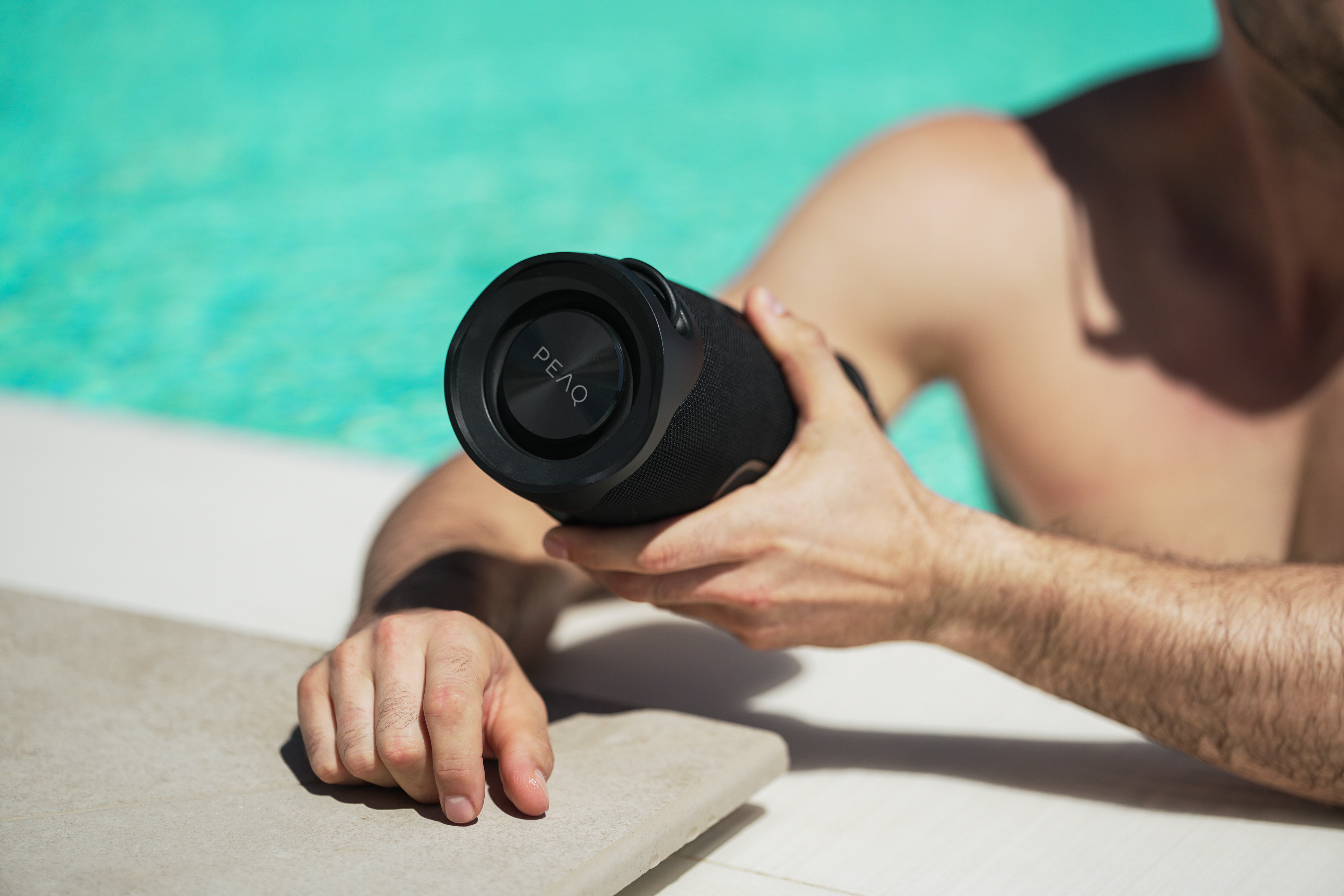 Eine Person hält einen schwarzen zylindrischen Lautsprecher in der Nähe eines Pools. Der Lautsprecher trägt das Wort PEAQ.