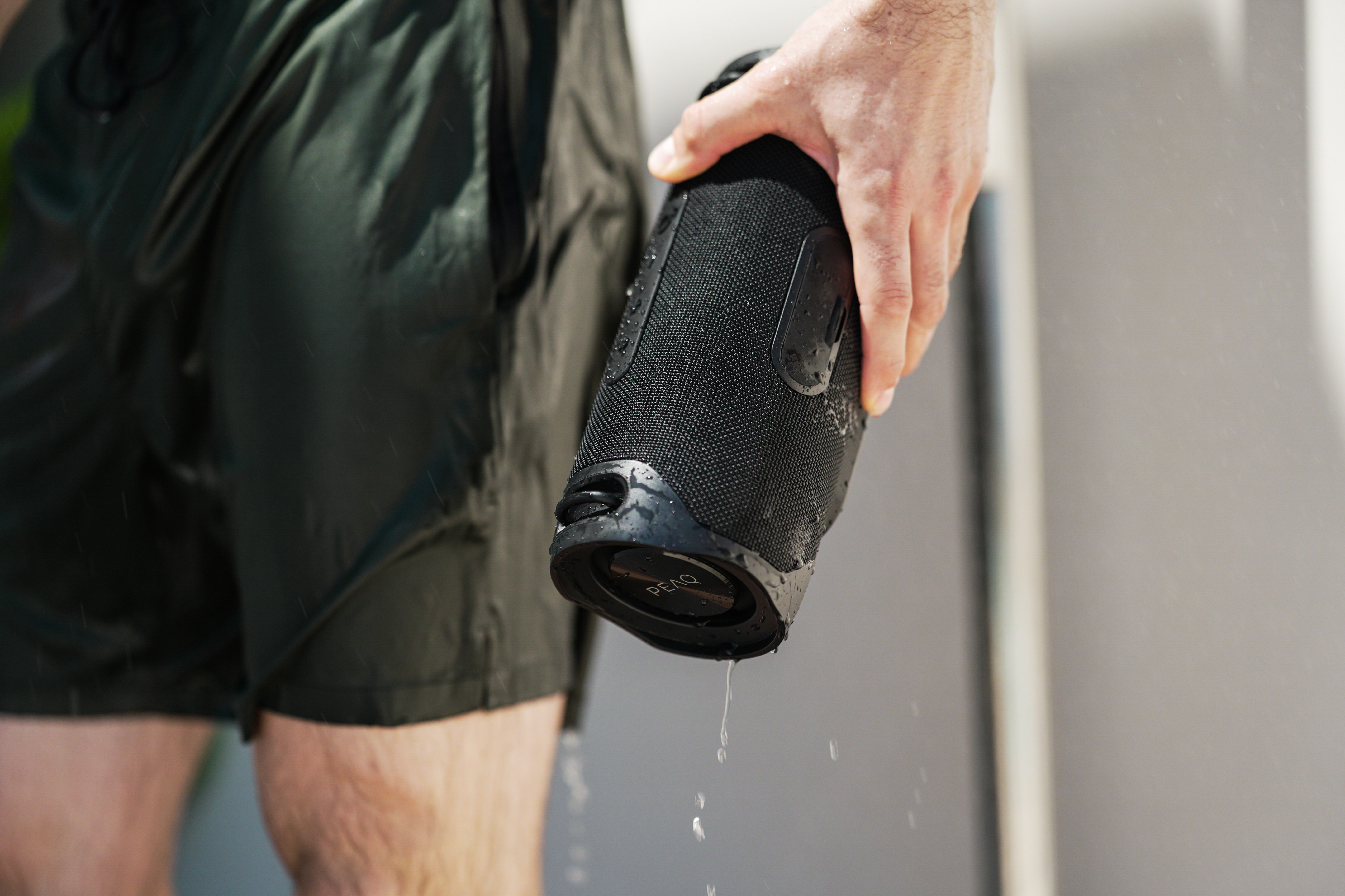 Eine Person hält einen schwarzen Lautsprecher mit Wassertropfen darauf. Die Person trägt schwarze Shorts. Wasser tropft.