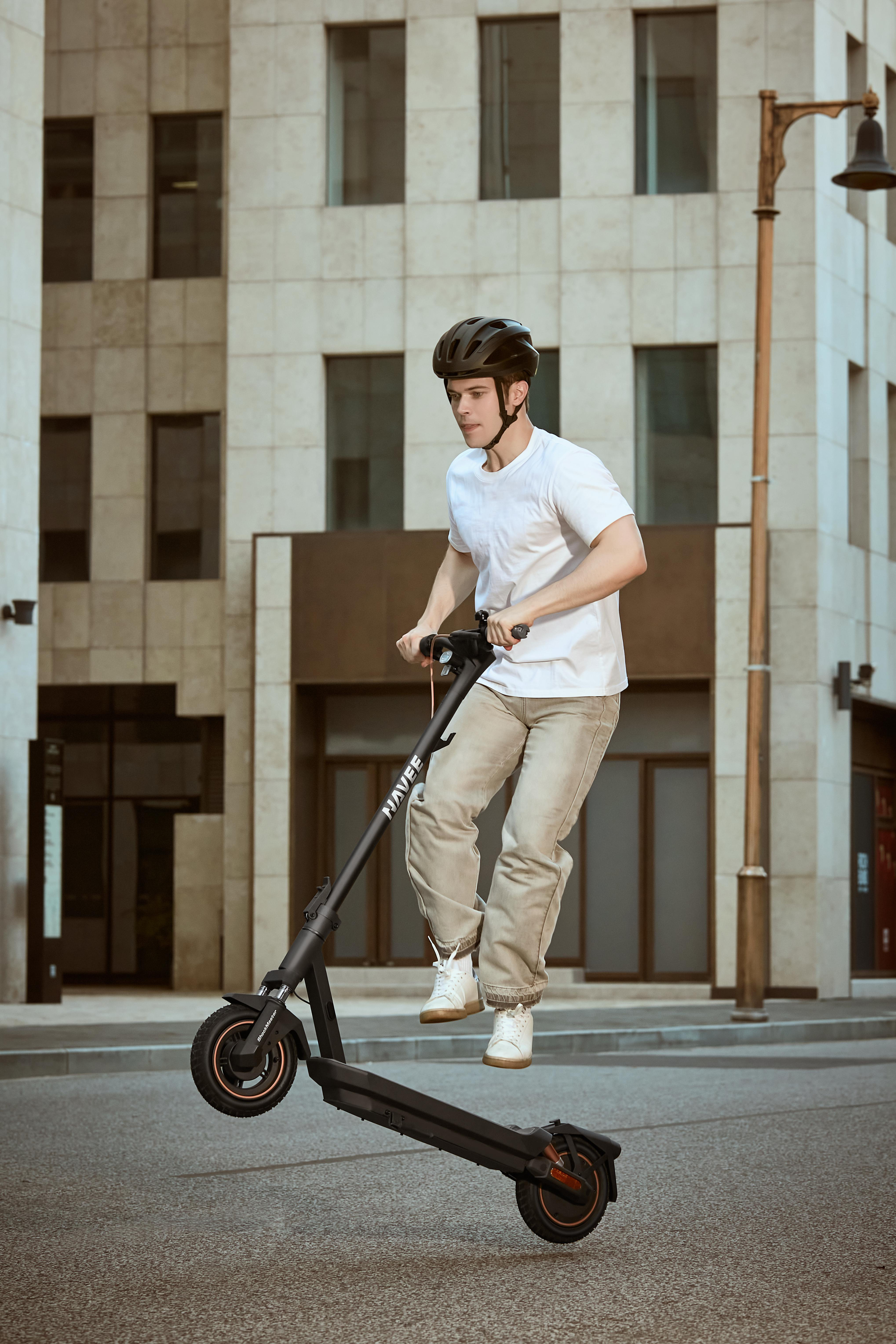 Ein Mann mit Helm springt mit einem Roller auf einer Stadtstraße. Gebäude im Hintergrund.