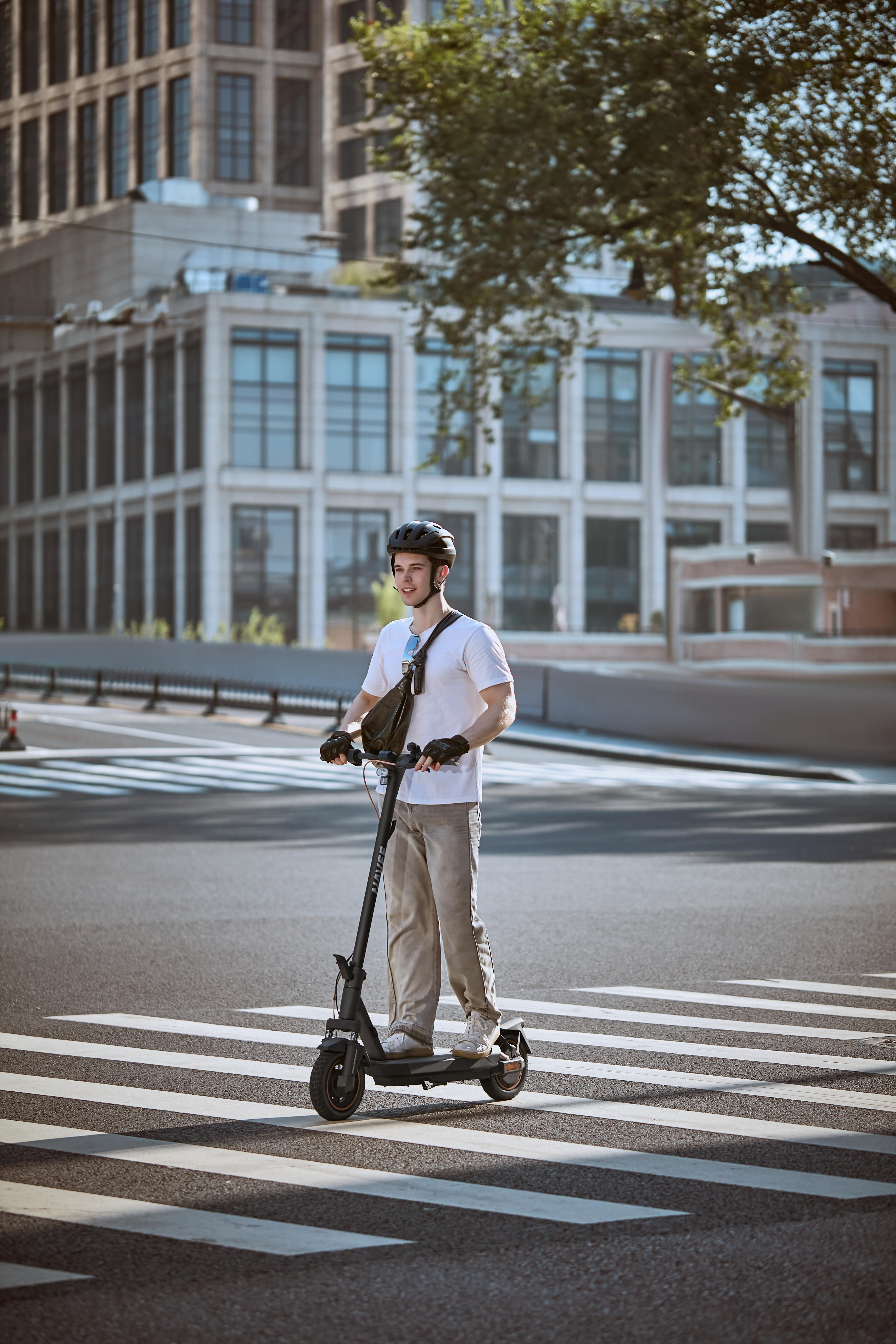 Ein Mann mit Helm fährt mit einem Roller auf einem Zebrastreifen. Gebäude und Bäume im Hintergrund.
