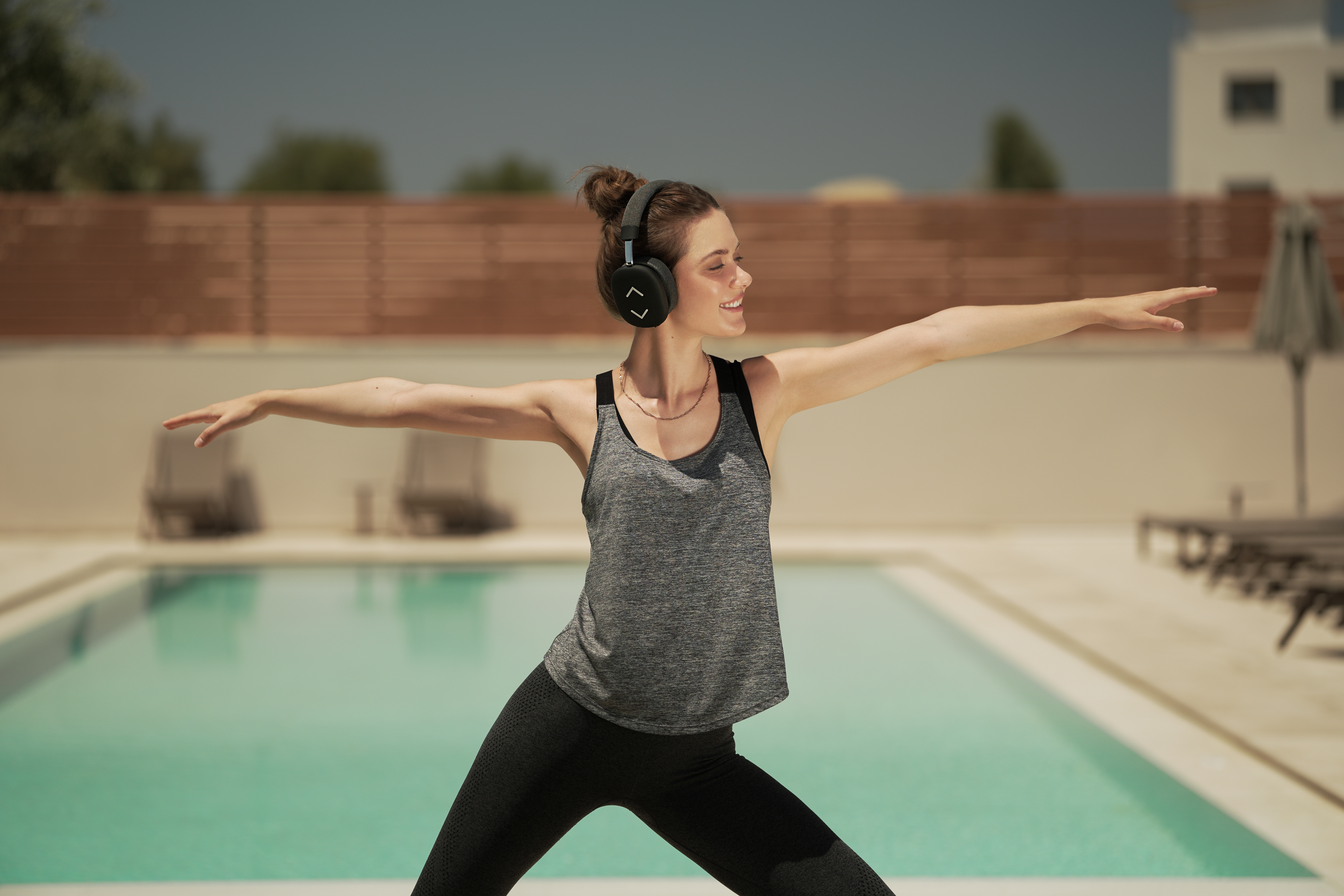 Eine Frau in Sportkleidung macht Yoga in der Nähe eines Pools. Sie trägt Kopfhörer und lächelt. Der Hintergrund hat einen Pool und einen Zaun.