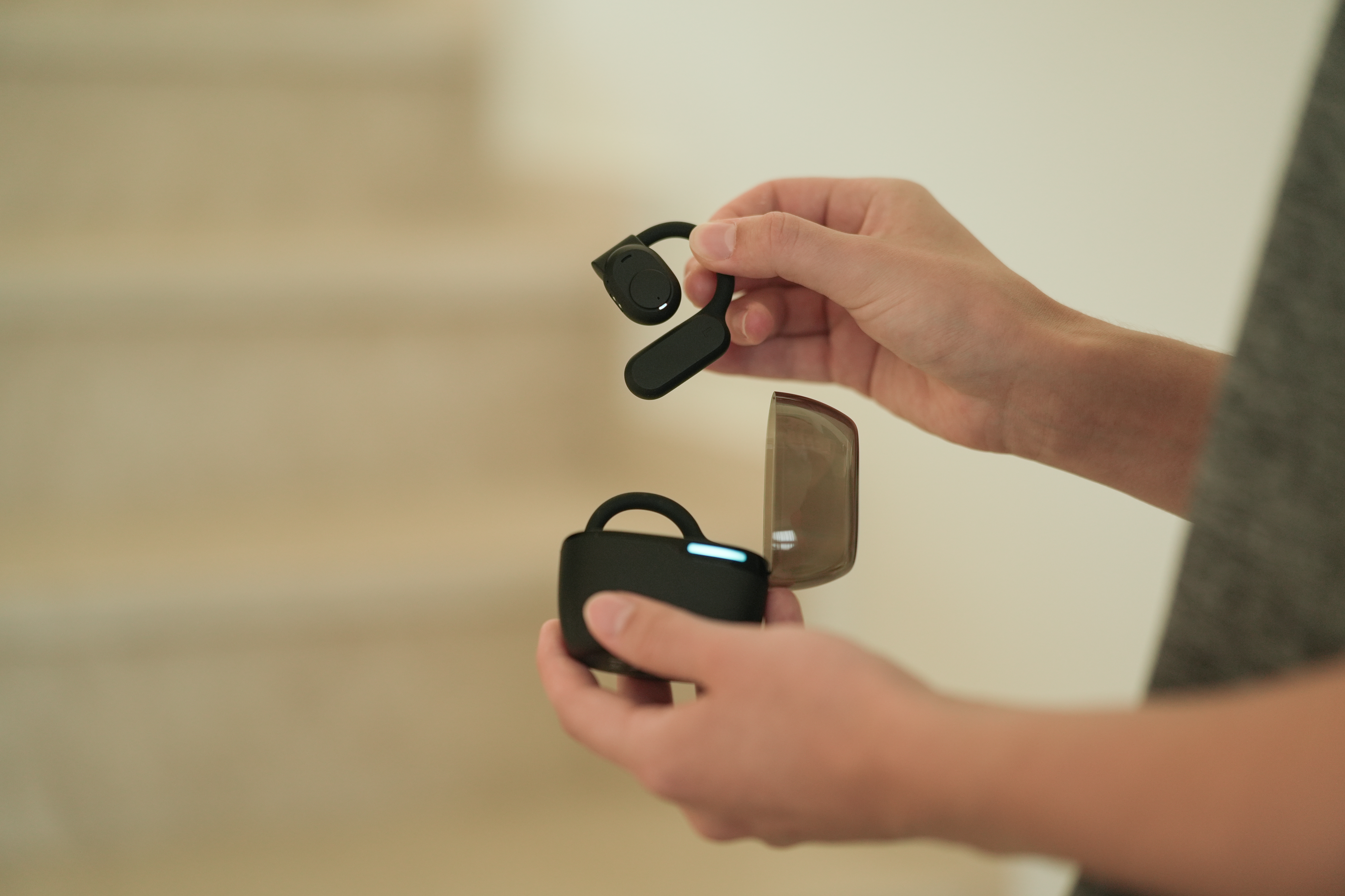 Eine Person hält ein schwarzes Headset und sein Ladeetui. Das Etui ist geöffnet. Der Hintergrund ist eine verschwommene beige Wand.