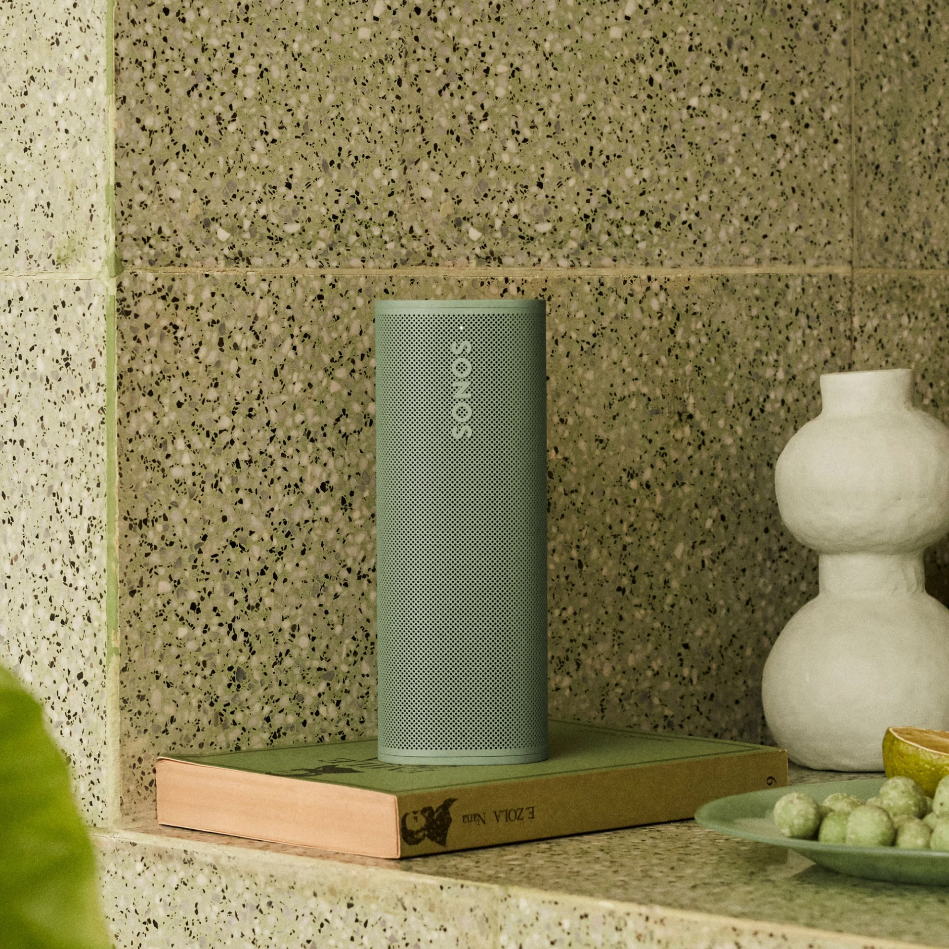 Zöld Sonos hangszóró egy könyvön, egy fehér váza mellett, egy zöld rágcsáló táljával és egy fél lime-mal.