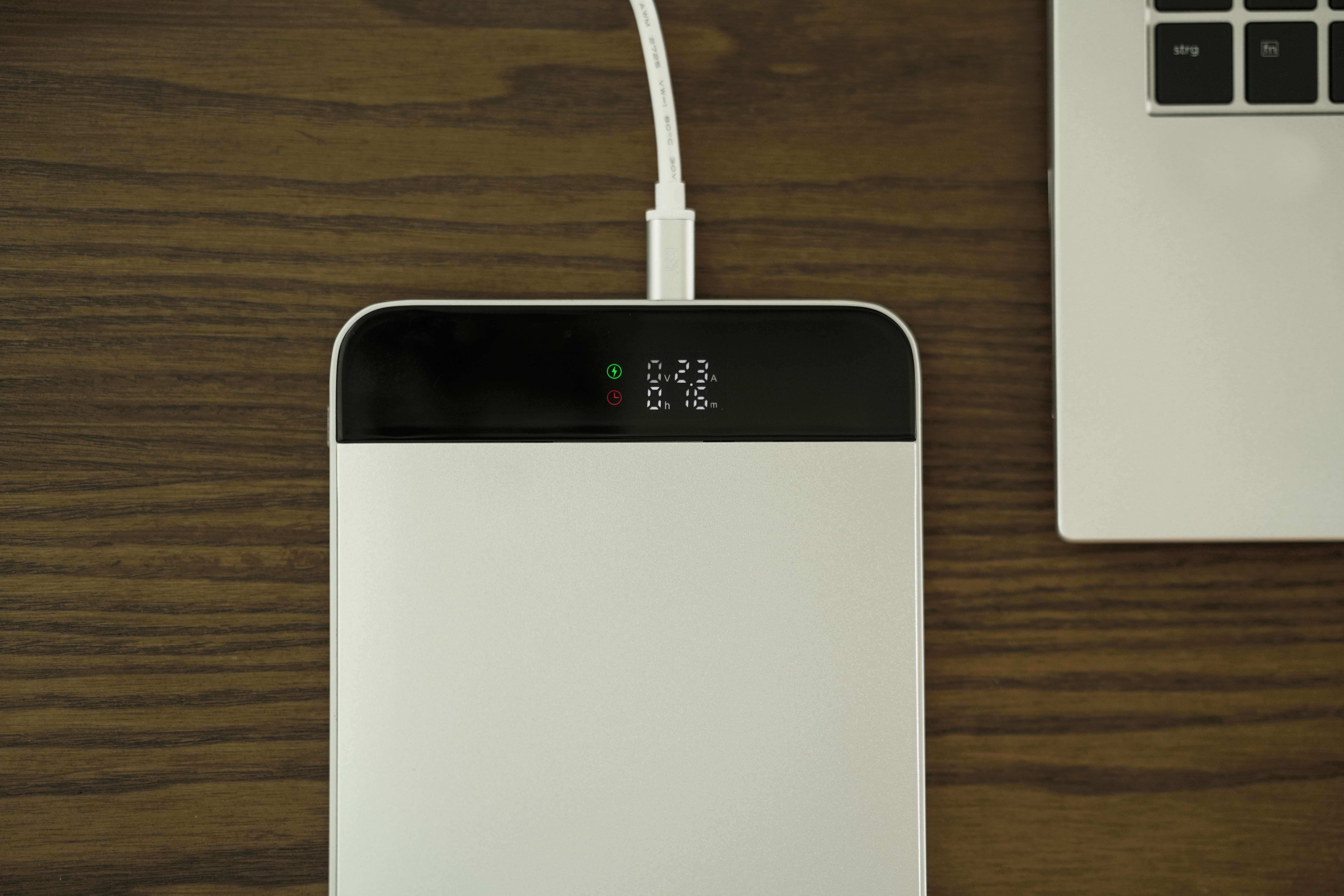 Eine silberne Powerbank wird aufgeladen, verbunden mit einem Laptop auf einem Holztisch. Der Bildschirm zeigt Ladeinfo.