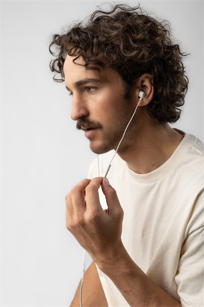 Egy göndör hajú férfi fülhallgatót visel, a kábelt tartja. Bajusza, orrkarikája és fehér pólója van, fehér háttér előtt.