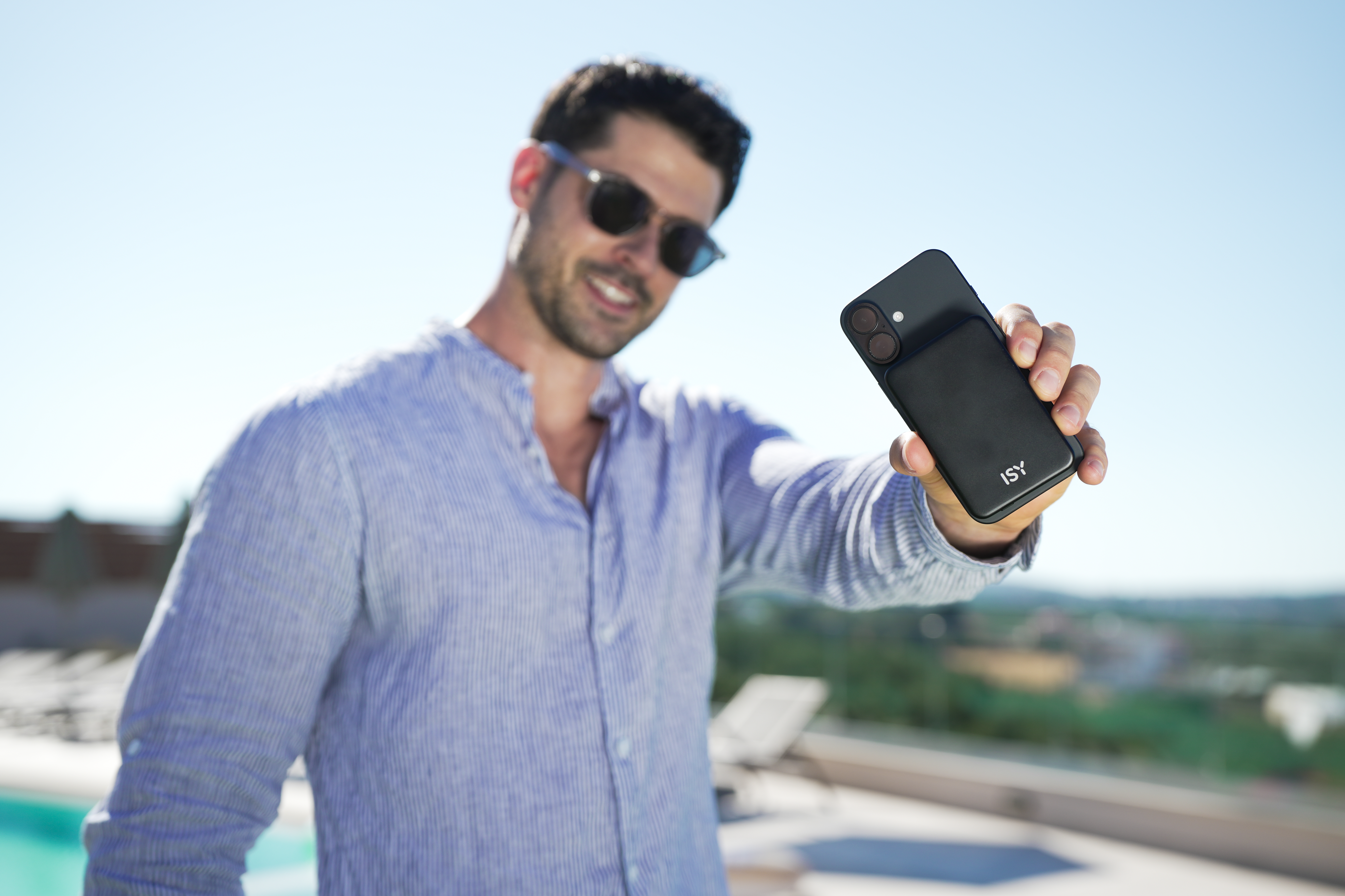 Mann mit Sonnenbrille hält ein schwarzes Telefon mit schwarzer Hülle hoch. Blauer Himmel im Hintergrund. Er lächelt.