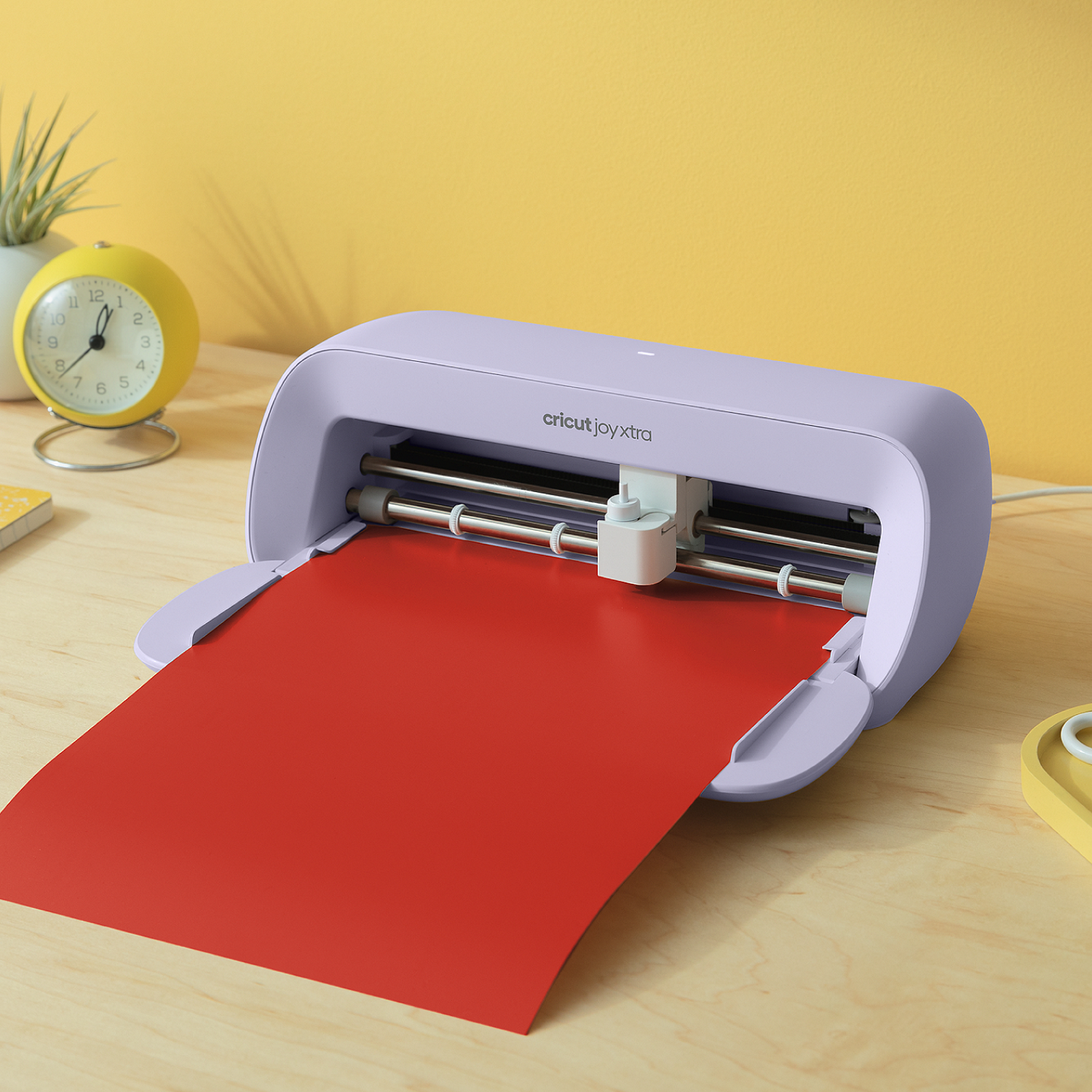 Eine Cricut Joy Xtra Maschine mit rotem Papier, das aus ihr herausragt. Links befinden sich eine gelbe Uhr und eine Pflanze. Der Hintergrund ist gelb.