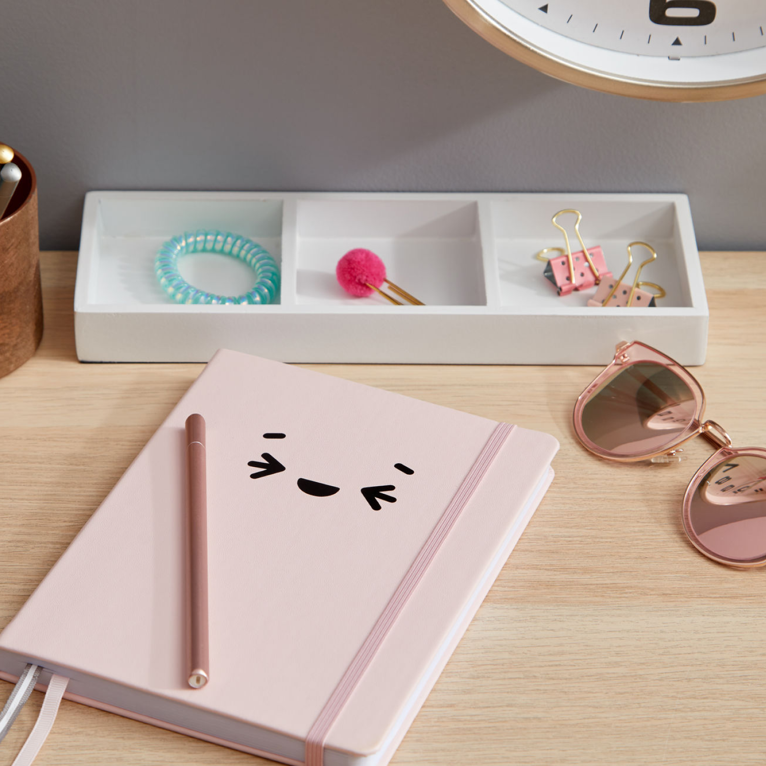 Rosa Notizbuch mit Gesicht, Stift, Sonnenbrille, Schreibtisch-Organizer mit Gegenständen und Uhr auf einem Holzschreibtisch.