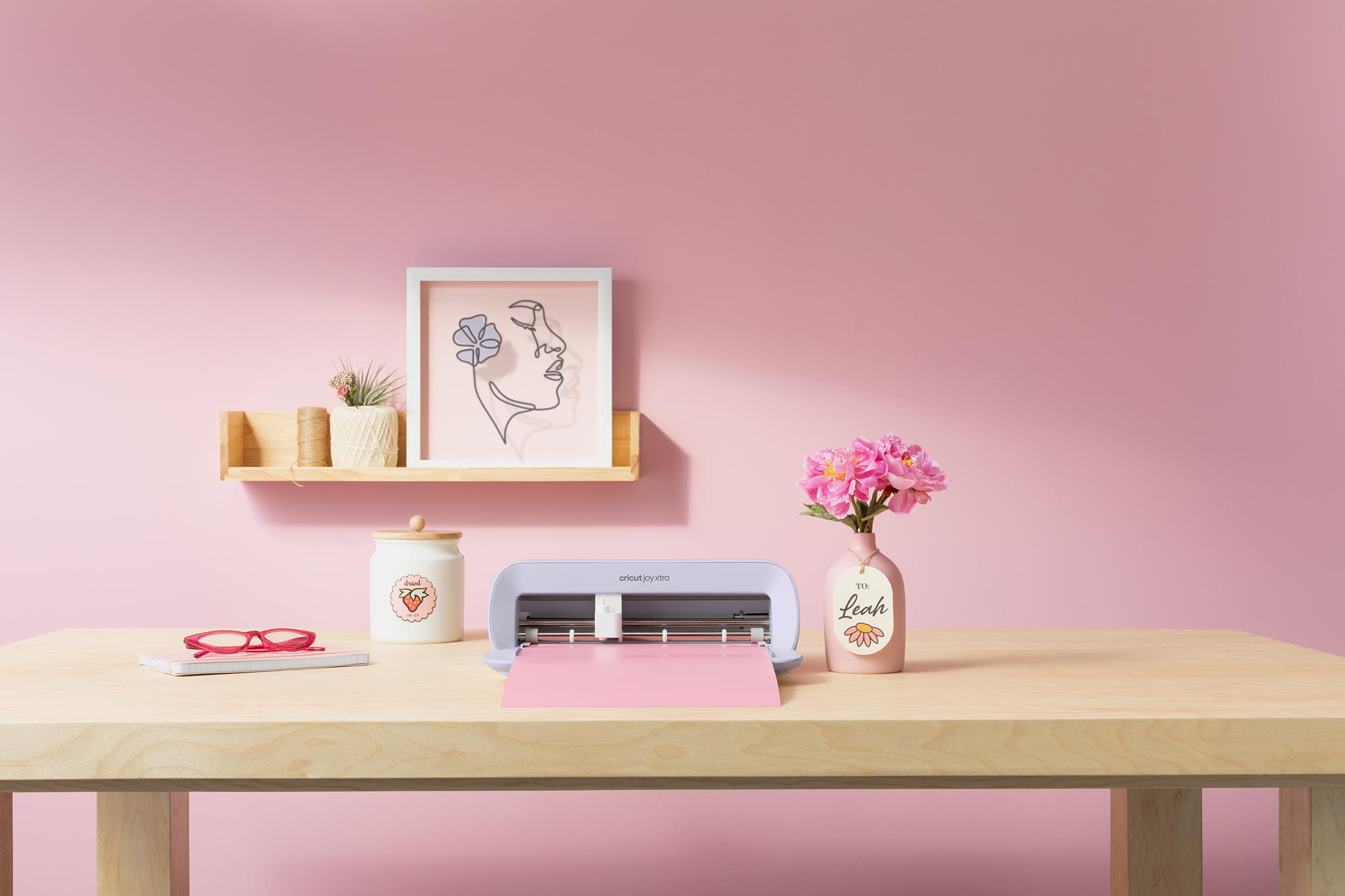 Ein Holztisch mit einer Cricut-Maschine, Blumen und Kunst vor rosa Hintergrund. Ein Schild sagt "To Leah".