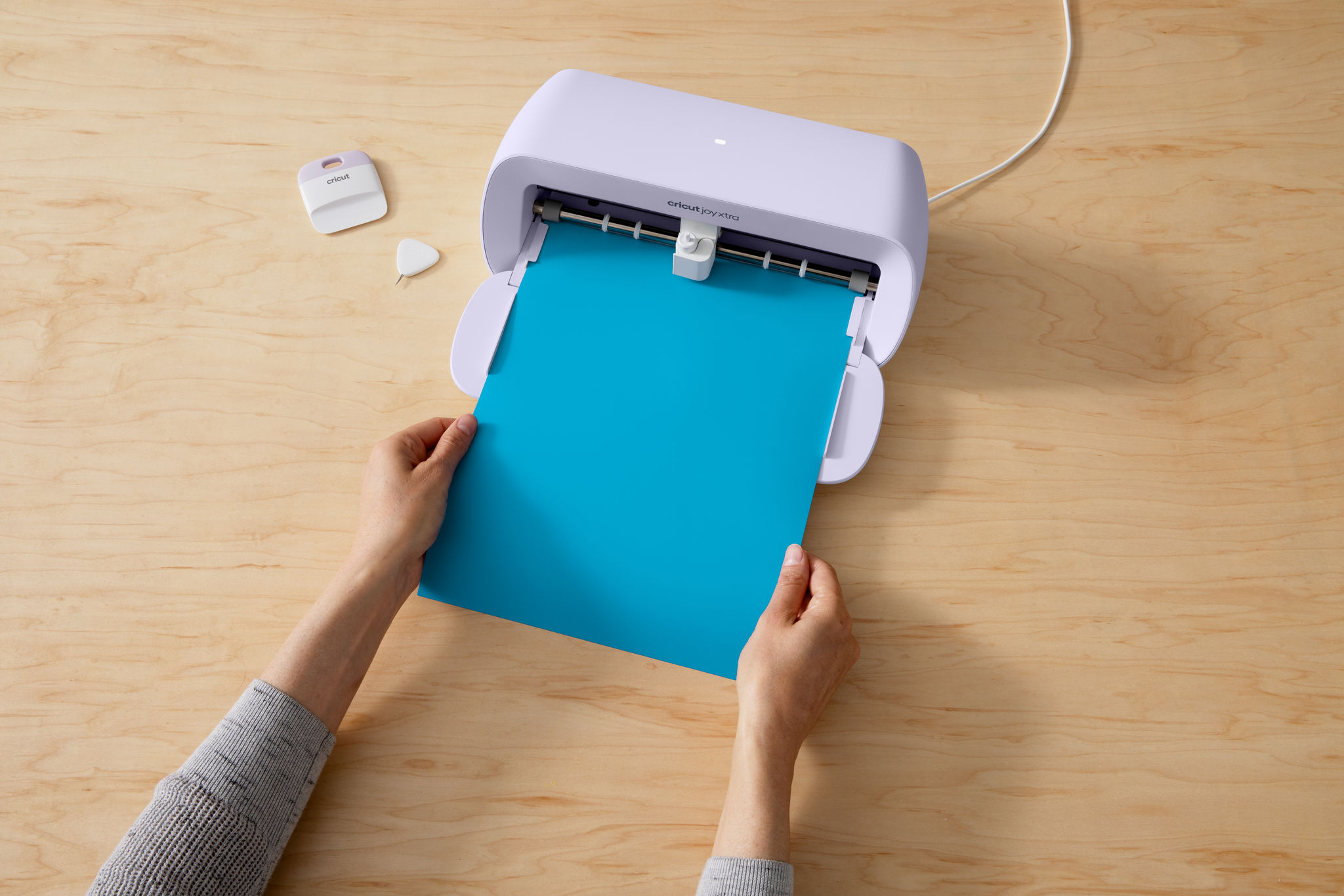 Hände halten ein blaues Papierblatt in einer Cricut-Maschine. Schaber und Entgittern Werkzeuge liegen auf einem Holztisch.