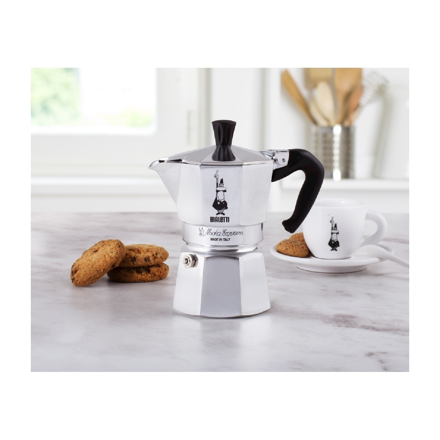 Una caffettiera Bialetti Moka Express argentata, tazza, piattino e biscotti su una superficie di marmo. Un cucchiaio e utensili da cucina sullo sfondo.
