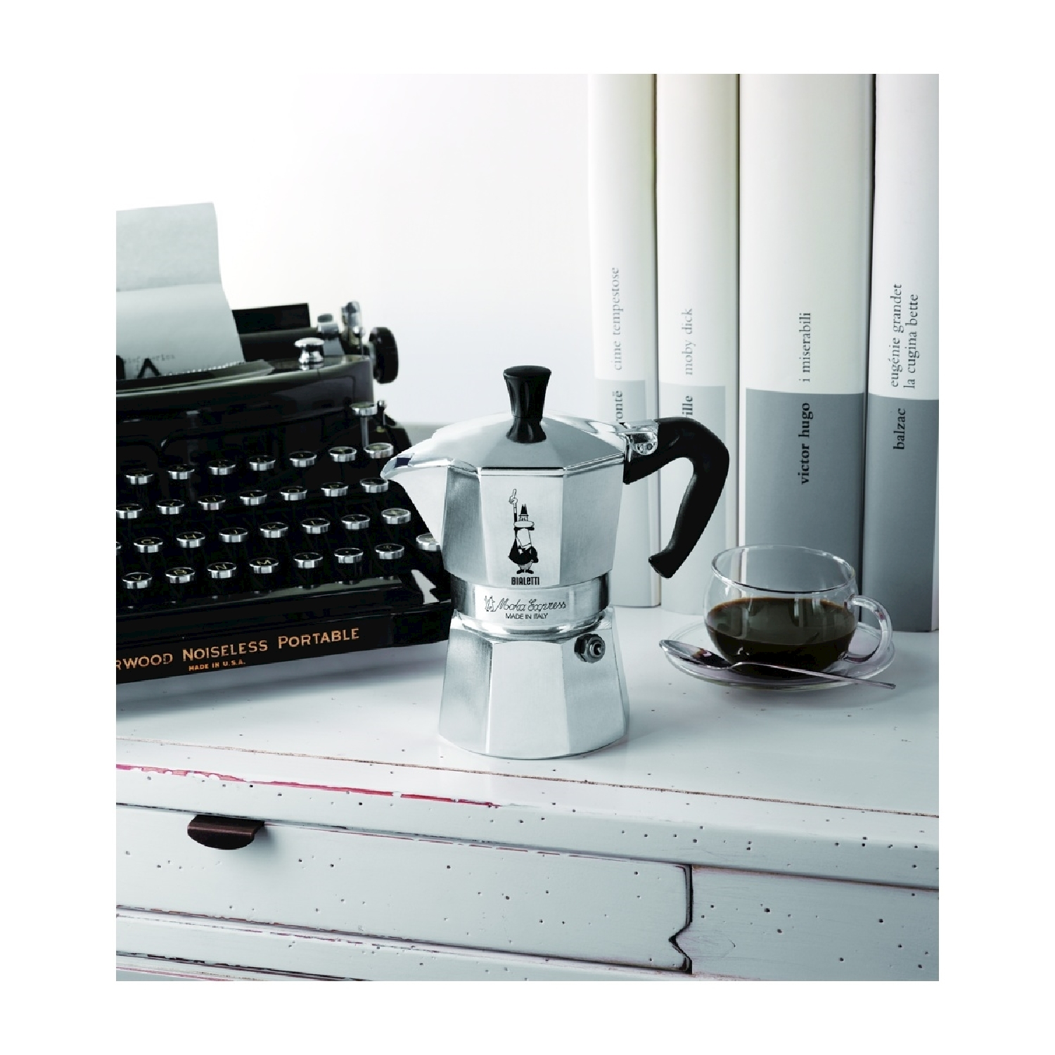 Una macchina da scrivere nera, una caffettiera d'argento, una tazza di caffè e libri su un tavolo bianco. La scena è ben illuminata.