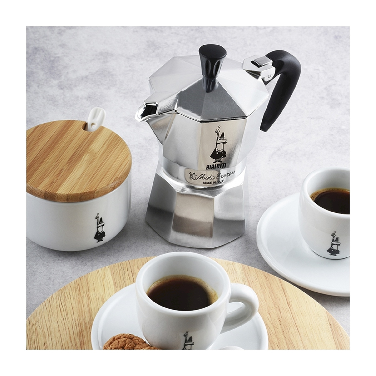 Una caffettiera Bialetti argentata, due tazzine di caffè espresso, una zuccheriera e un tagliere in legno su una superficie grigio chiaro.