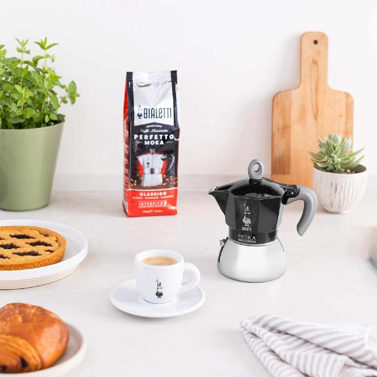 Una scena di cucina con caffè, moka, pasticcini e piante. Una busta di caffè è dietro una tazzina di caffè.