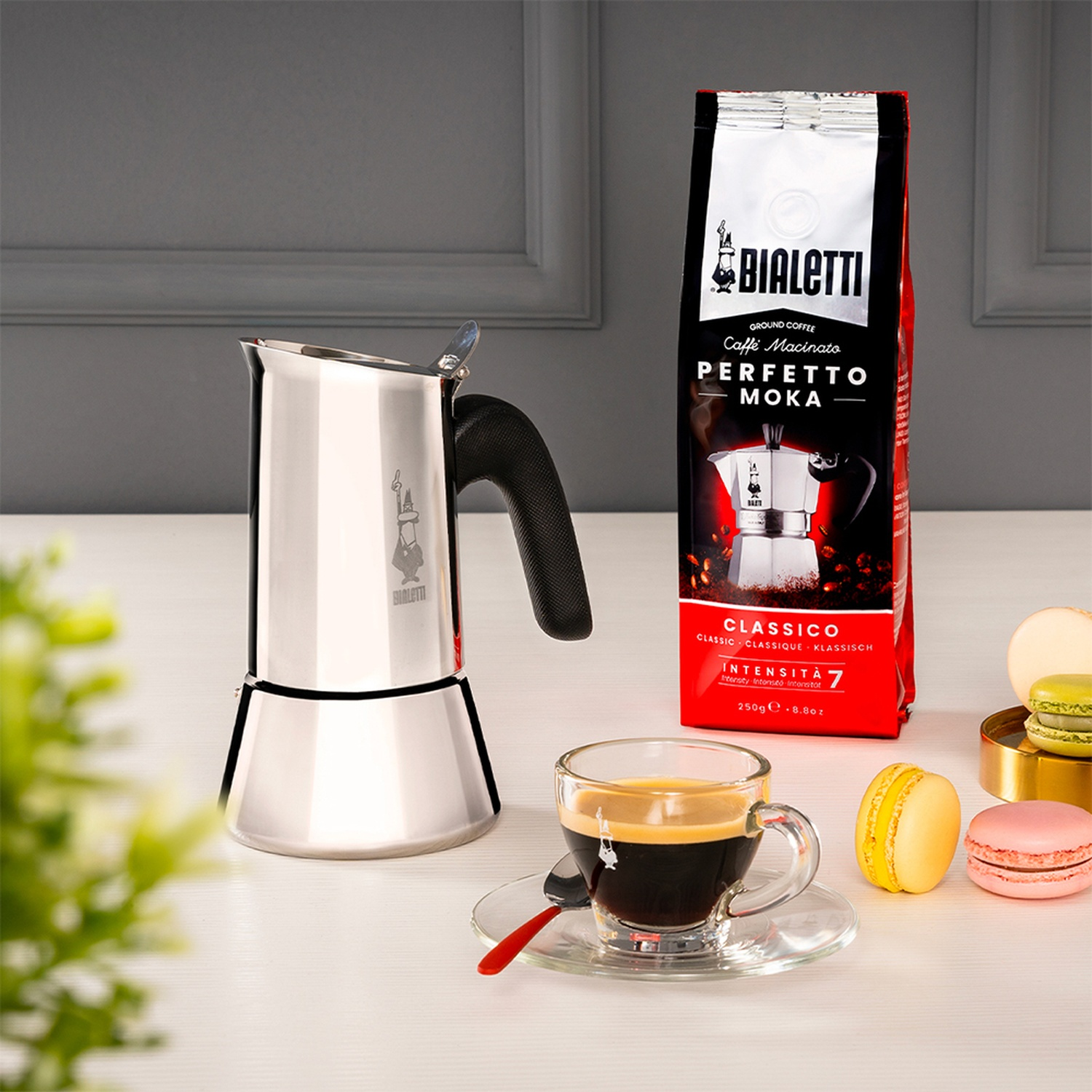 Una natura morta con una caffettiera moka Bialetti, caffè, una confezione di caffè e macaron su un tavolo. Lo sfondo è un muro grigio.
