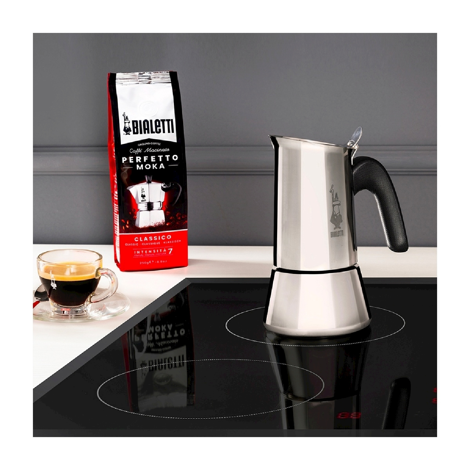 Un'attrezzatura per il caffè con una caffettiera moka Bialetti su un fornello, un sacchetto di caffè e una tazza di caffè, il tutto su una superficie bianca.