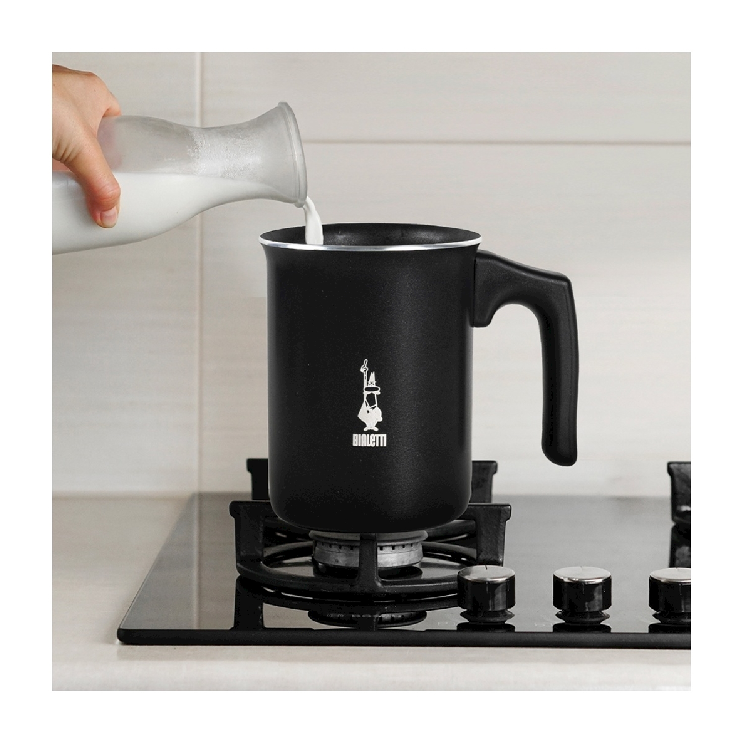 Una mano versa il latte da una bottiglia di vetro in una pentola nera su un piano cottura. La pentola ha un manico e il logo Bialetti.