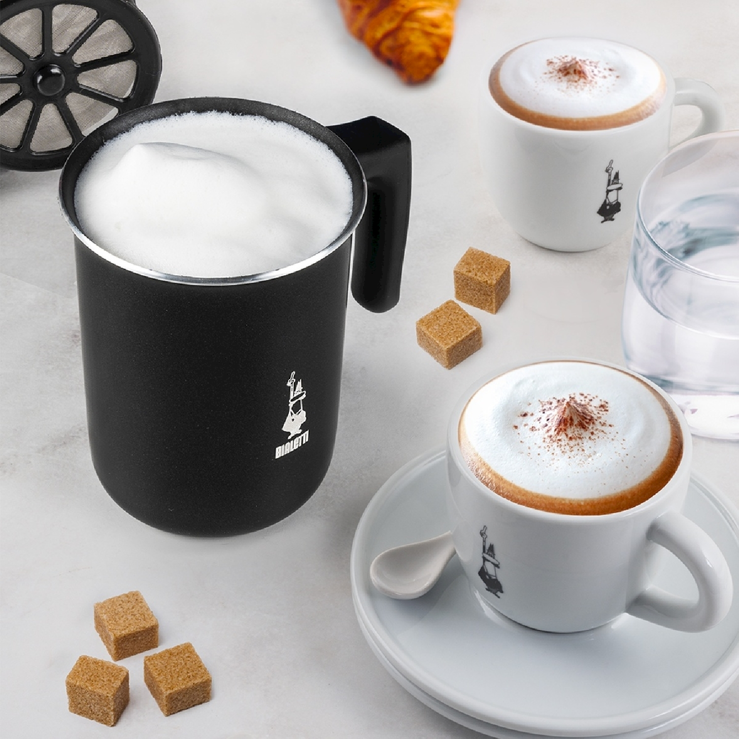 Una natura morta con cappuccino schiumoso, montalatte nero, zollette di zucchero e croissant su superficie bianca.