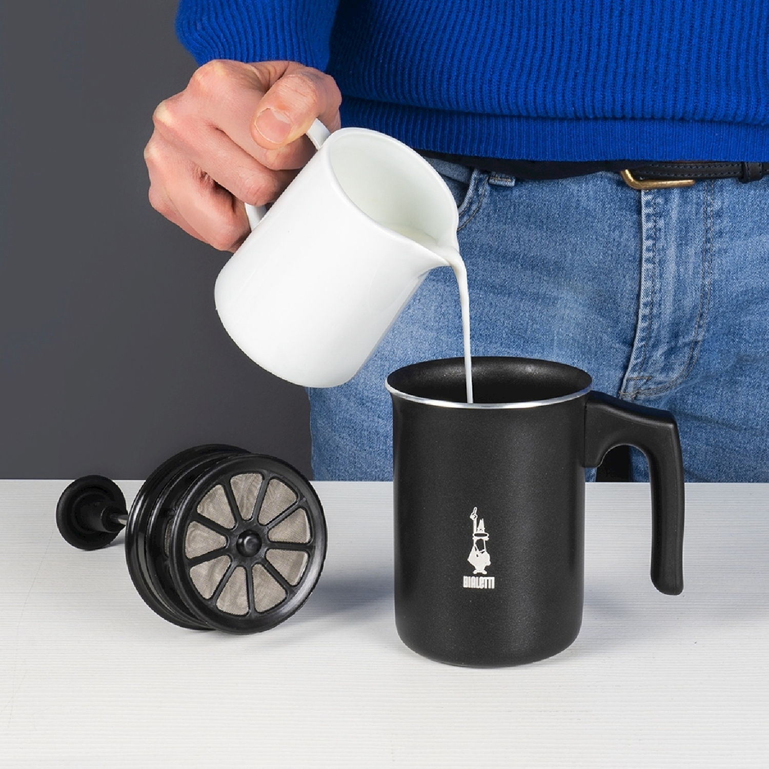 Una persona versa latte da una brocca bianca in una caffettiera nera. Una pressa francese è a sinistra. La persona indossa il blu.