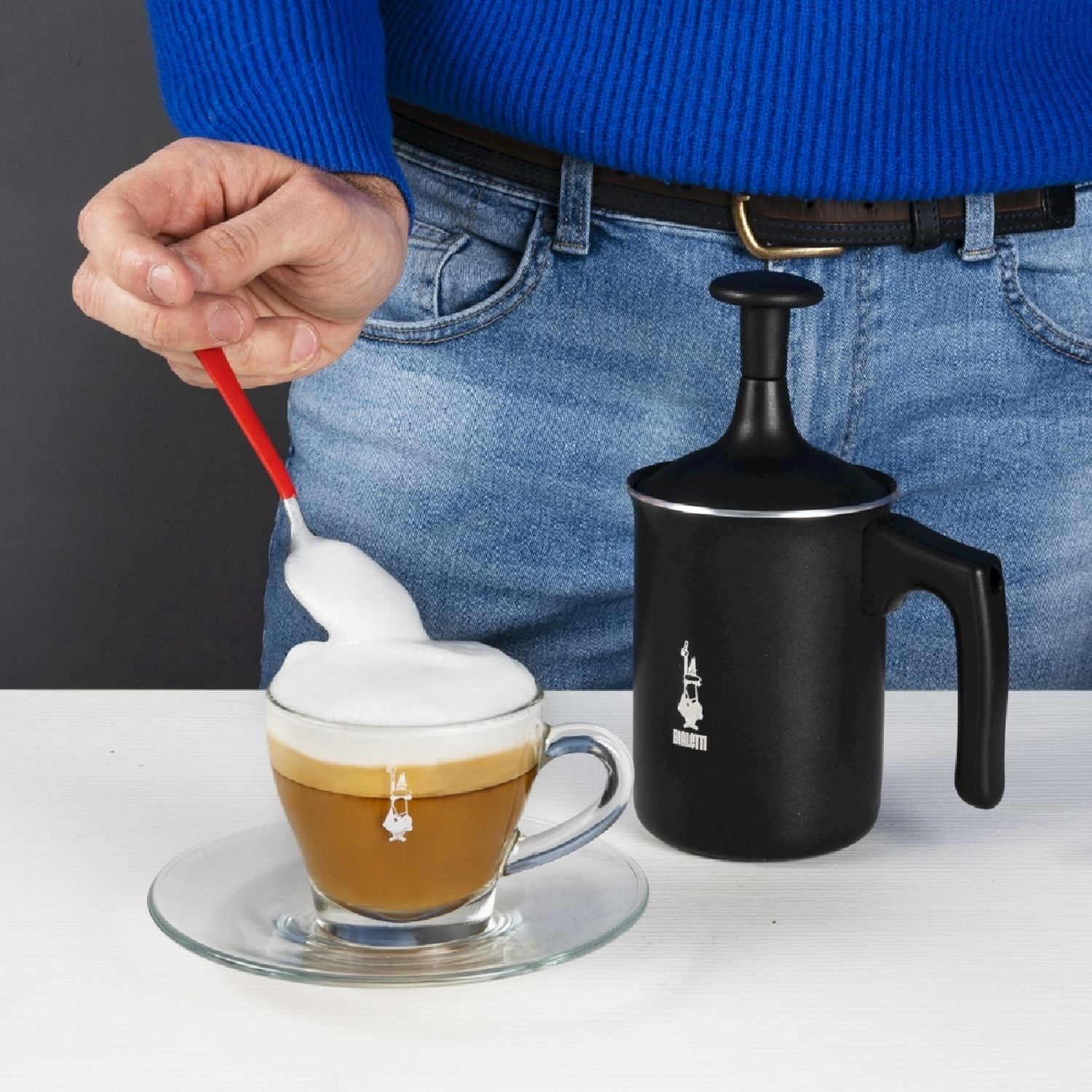 Una persona sta preparando il caffè con la schiuma. Una tazza di caffè con schiuma su un piattino. Accanto, una caffettiera nera.