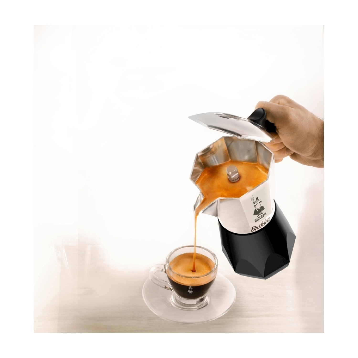 Una mano versa espresso da una caffettiera in una tazza di vetro. La caffettiera è argentata e nera. Il caffè è scuro e ricco.