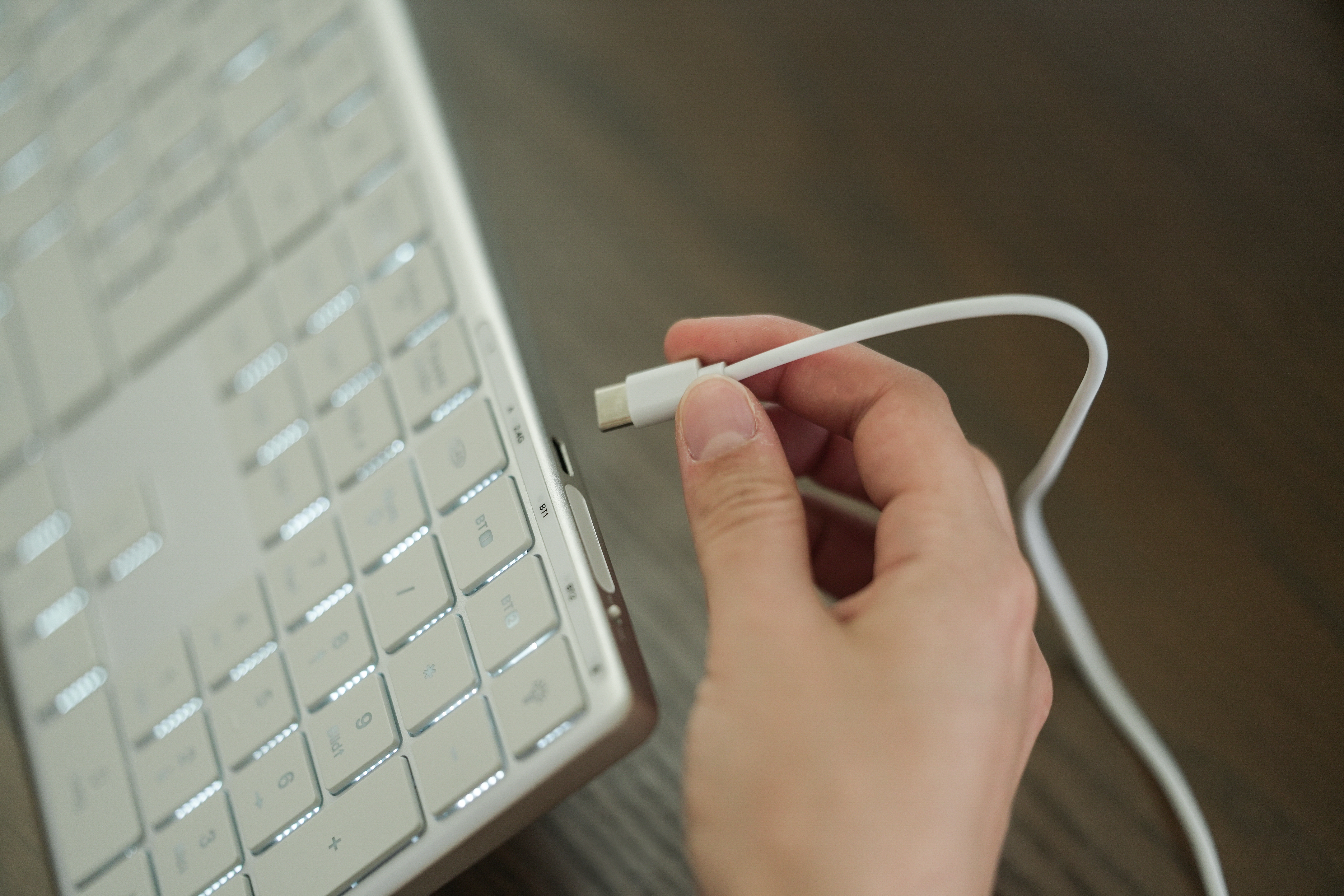 Hand hält weißes USB-C-Kabel, um es in eine weiße, hintergrundbeleuchtete Tastatur einzustecken. Braune Holzoberfläche.
