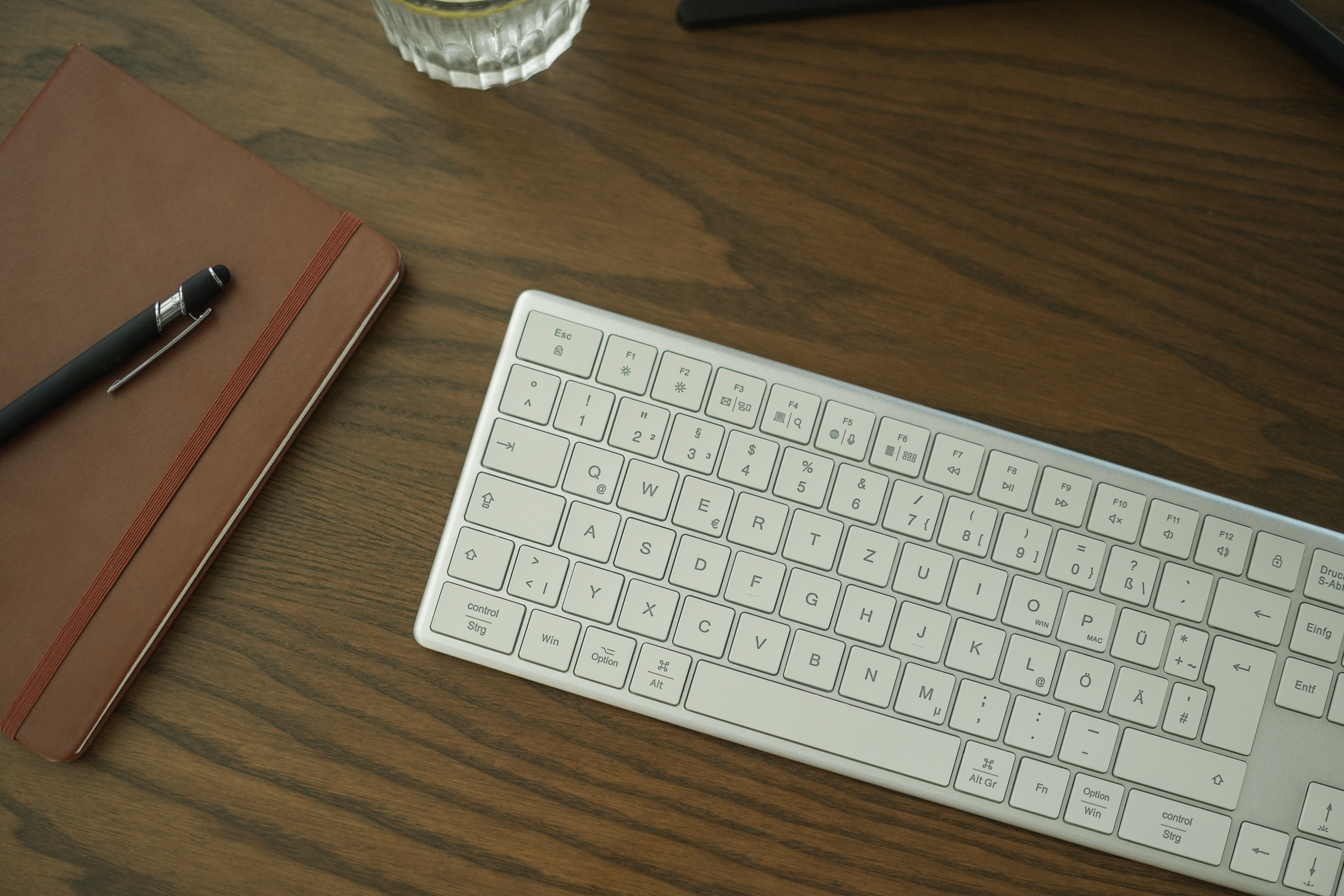 Arbeitsplatz mit Tastatur, Notizbuch und Stift. Alles auf einem Holzschreibtisch.