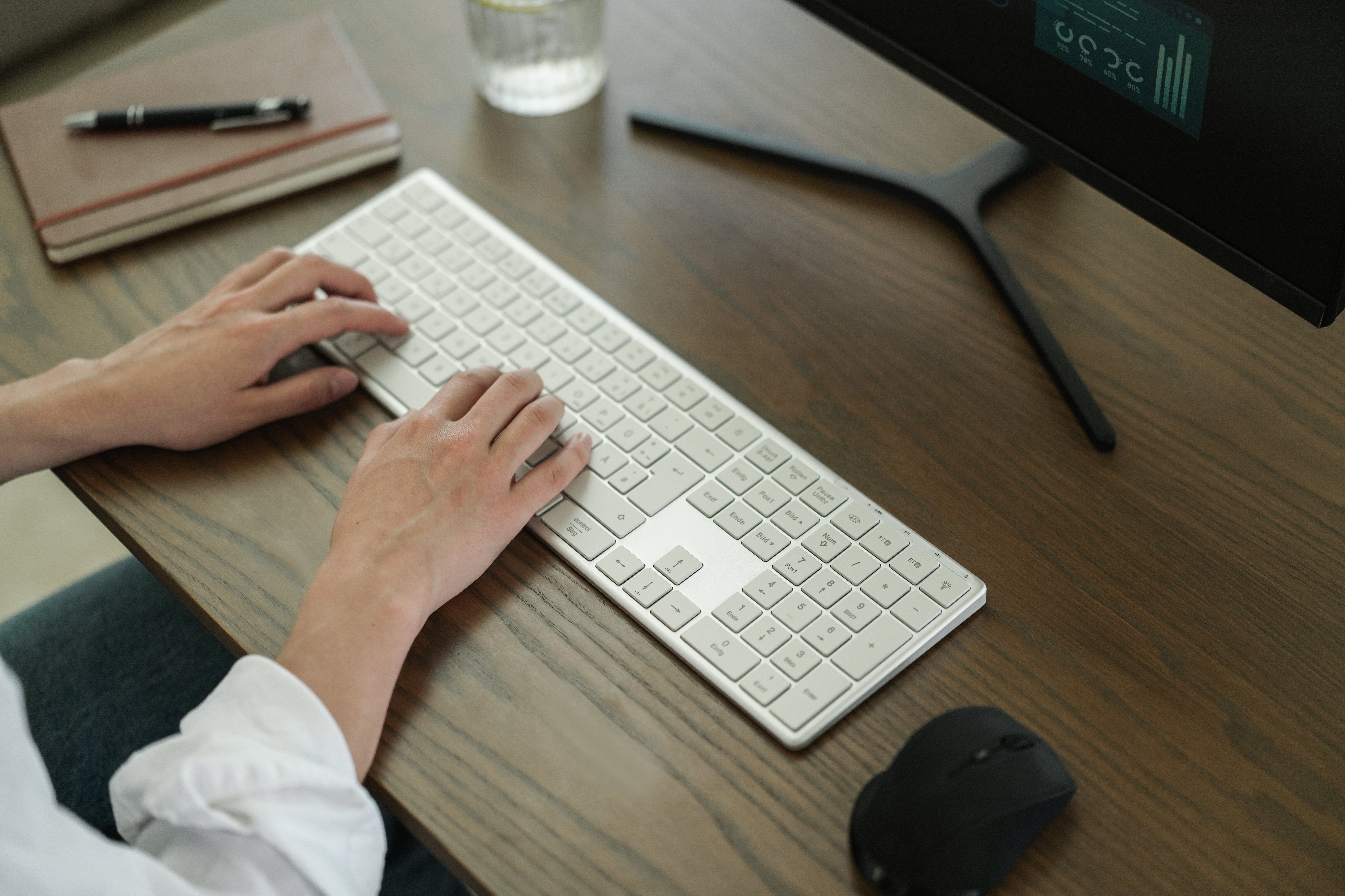 Hände tippen auf einer weißen Tastatur auf einem Holzschreibtisch. Ein Monitor, Maus und Notizbuch sind ebenfalls sichtbar.