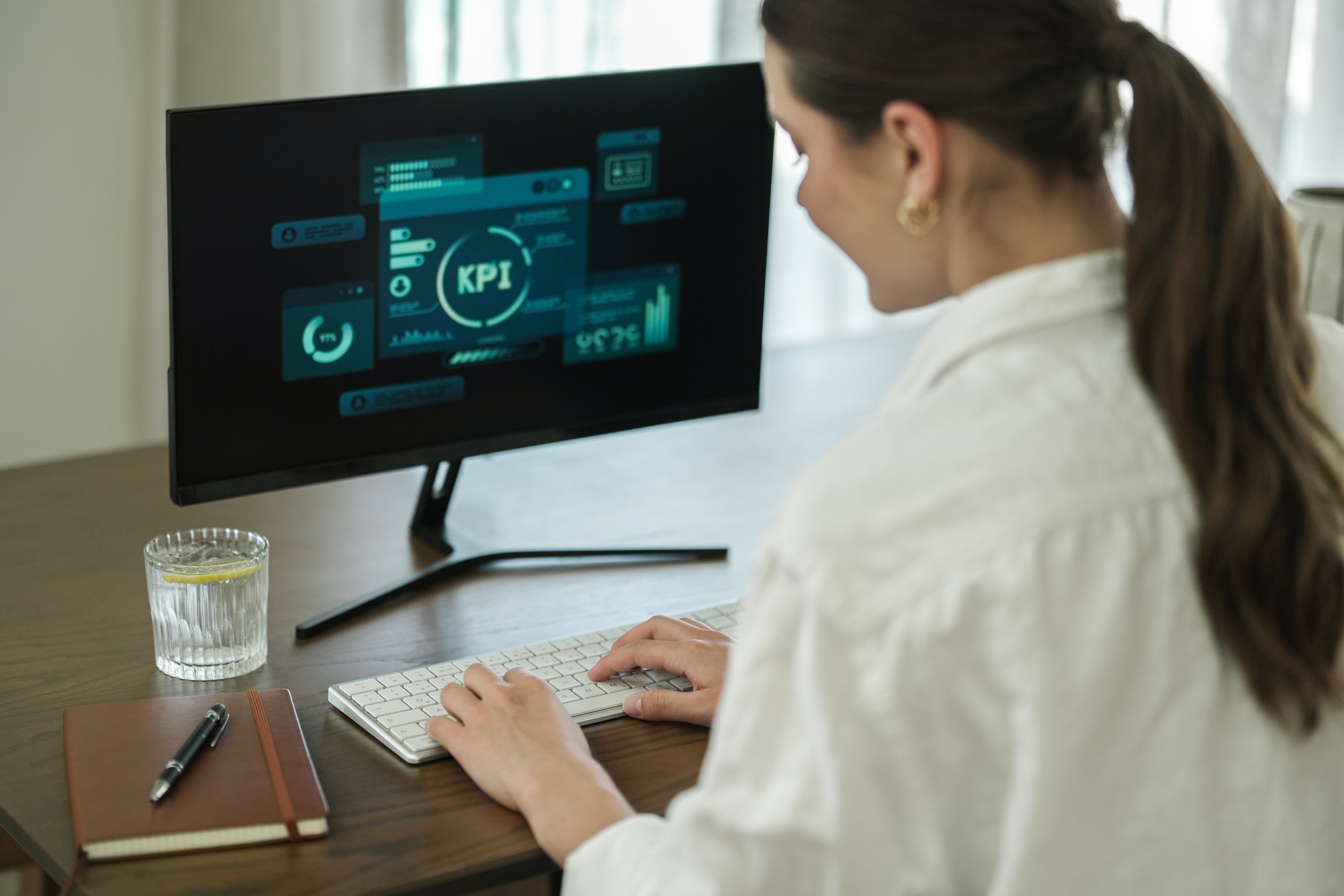 Frau tippt auf einer Tastatur, KPI-Daten auf einem Monitor. Glas Wasser und Notizbuch auf dem Tisch.