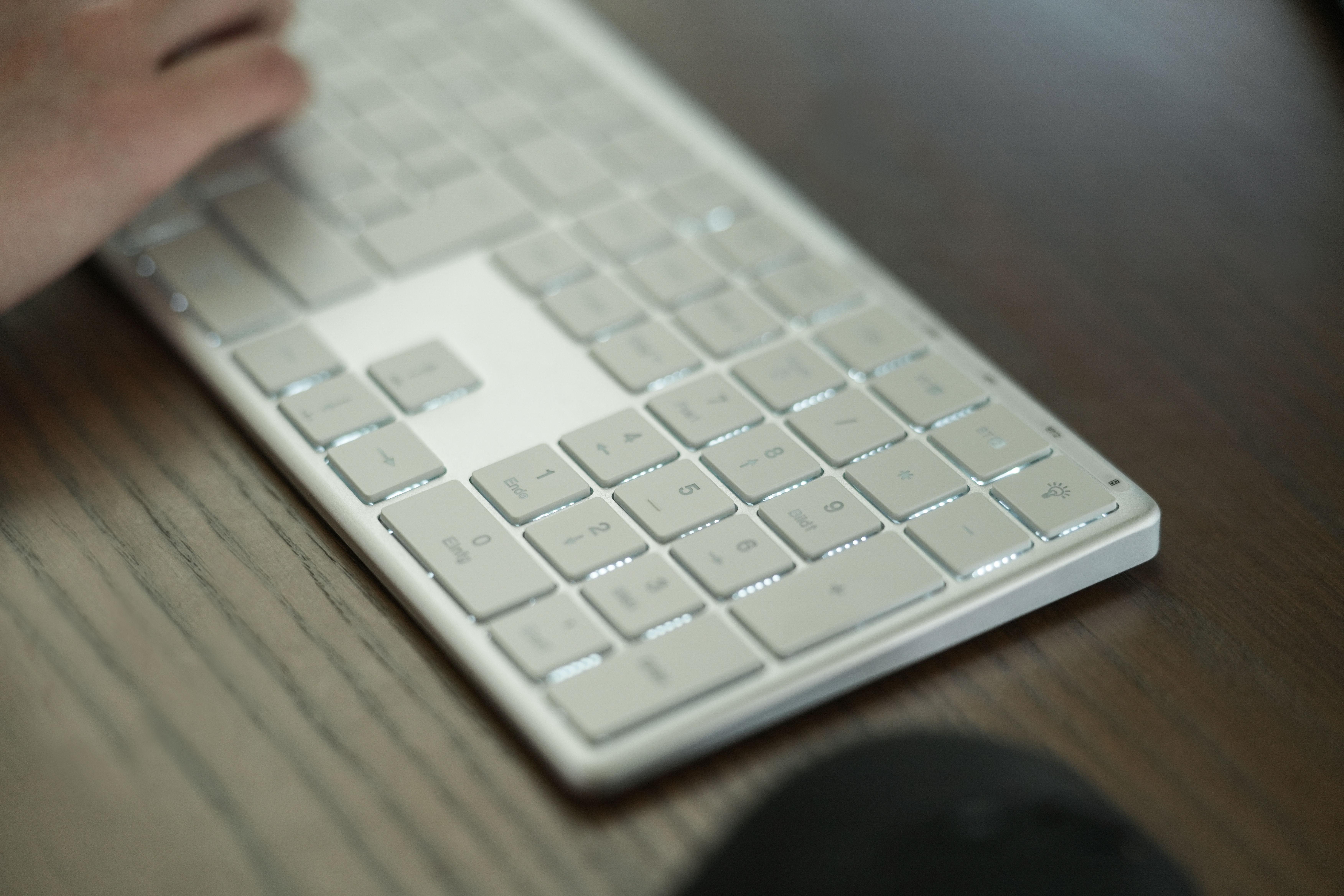 Eine weiße Tastatur mit beleuchteten Tasten liegt auf einem Holztisch. Eine Hand liegt auf der Tastatur, die Tasten sind im Fokus.