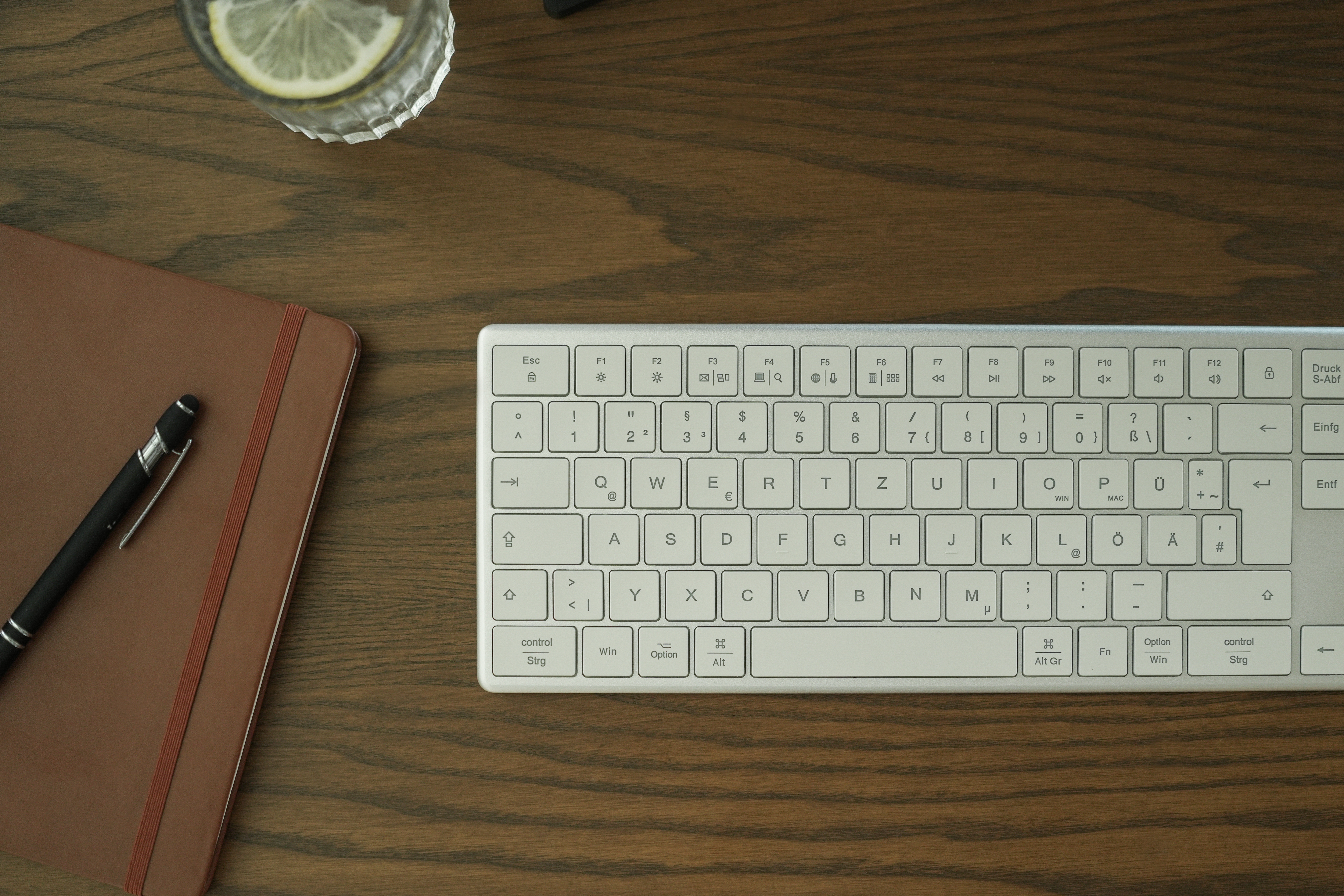 Eine Tastatur, ein Notizbuch, ein Stift und ein Glas Wasser mit Zitrone auf Holz.