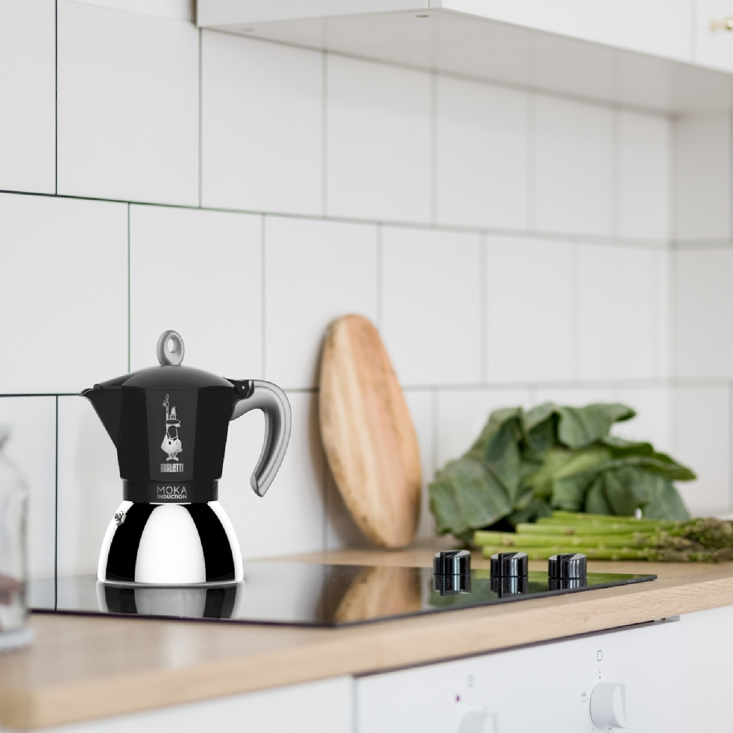 Caffettiera Moka nera e argento su un piano cottura nero. Tavola di legno, verdure verdi e parete piastrellata bianca sullo sfondo.