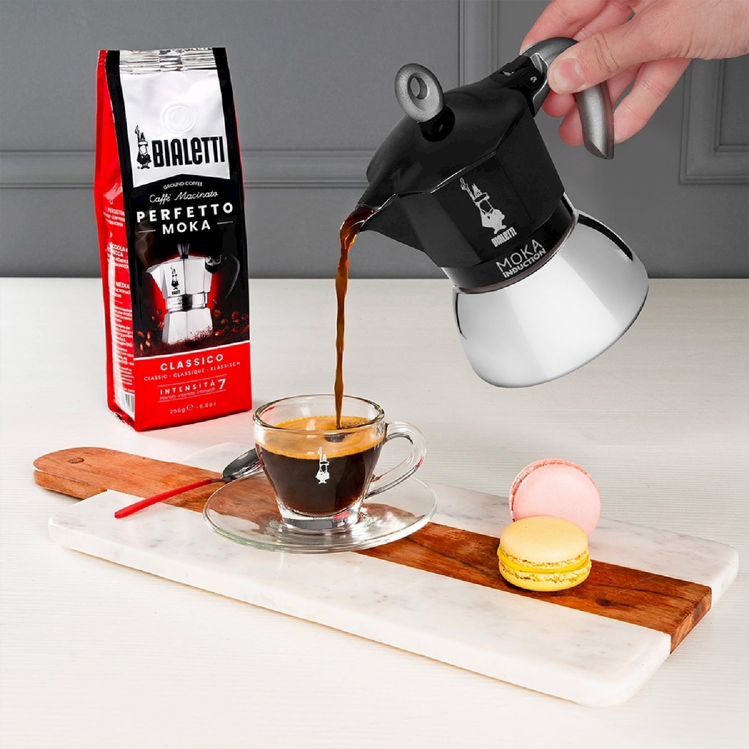 Una caffettiera moka nera versa il caffè in una tazza di vetro. Un sacchetto di caffè e macaron sono su una tavola di legno.