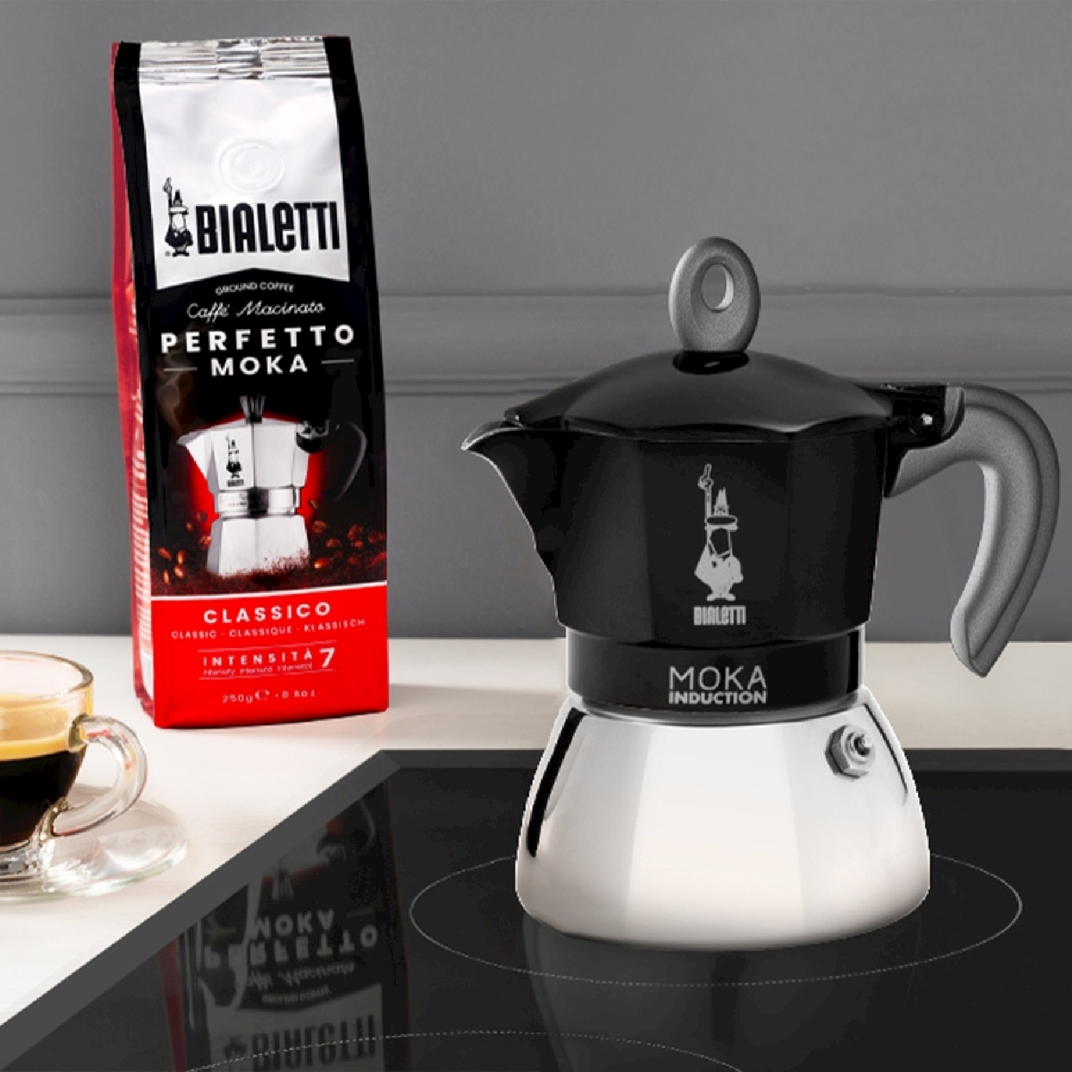 Una caffettiera Bialetti Moka accanto a un sacchetto di caffè. Una tazza di caffè a sinistra. La scena è su un piano cottura nero.
