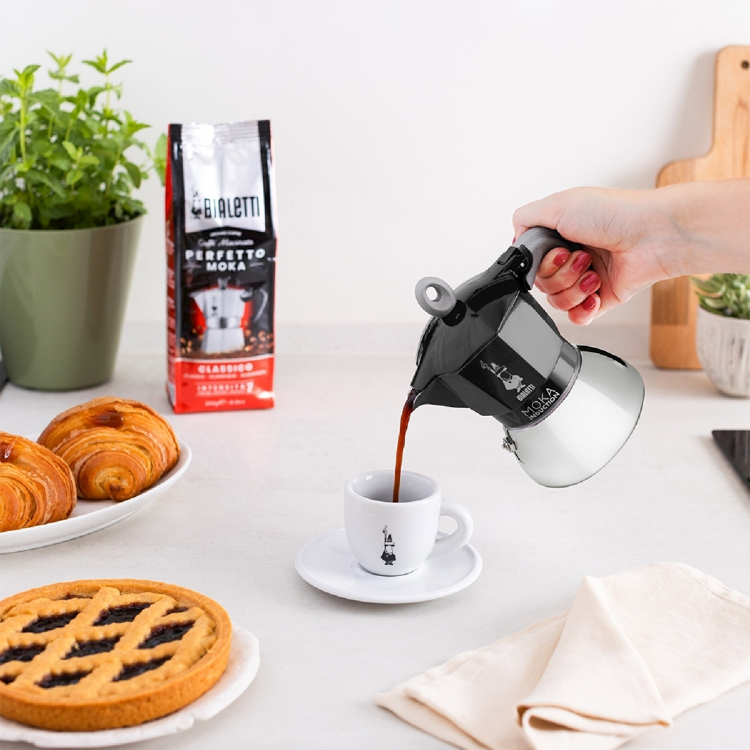 Una persona versa il caffè da una caffettiera moka nera e argento in una tazza bianca. Cornetti, una crostata e chicchi di caffè sono vicini.