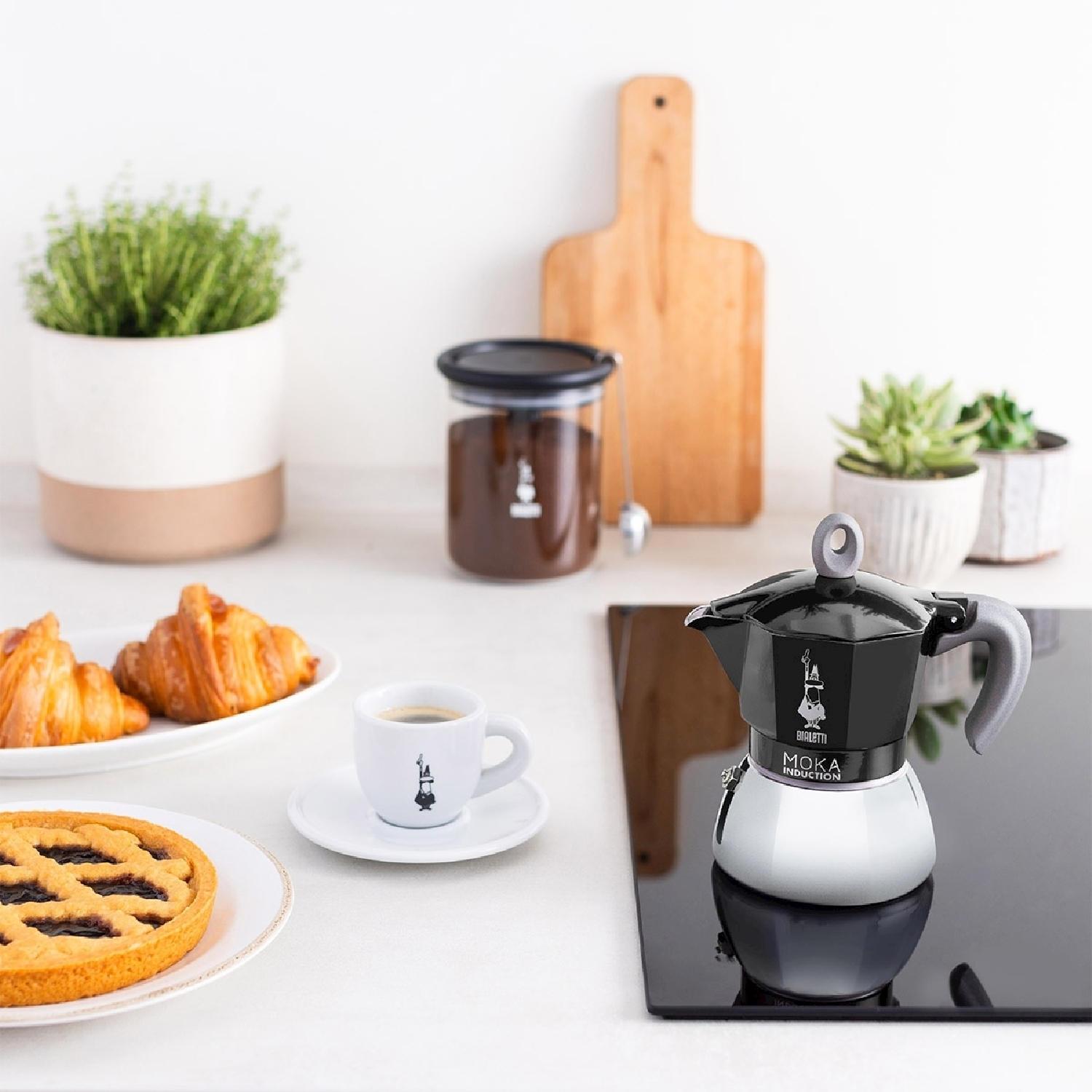 Una scena di cucina con caffettiera, tazza, croissant e torta. Ci sono piante e un barattolo di chicchi di caffè sullo sfondo.