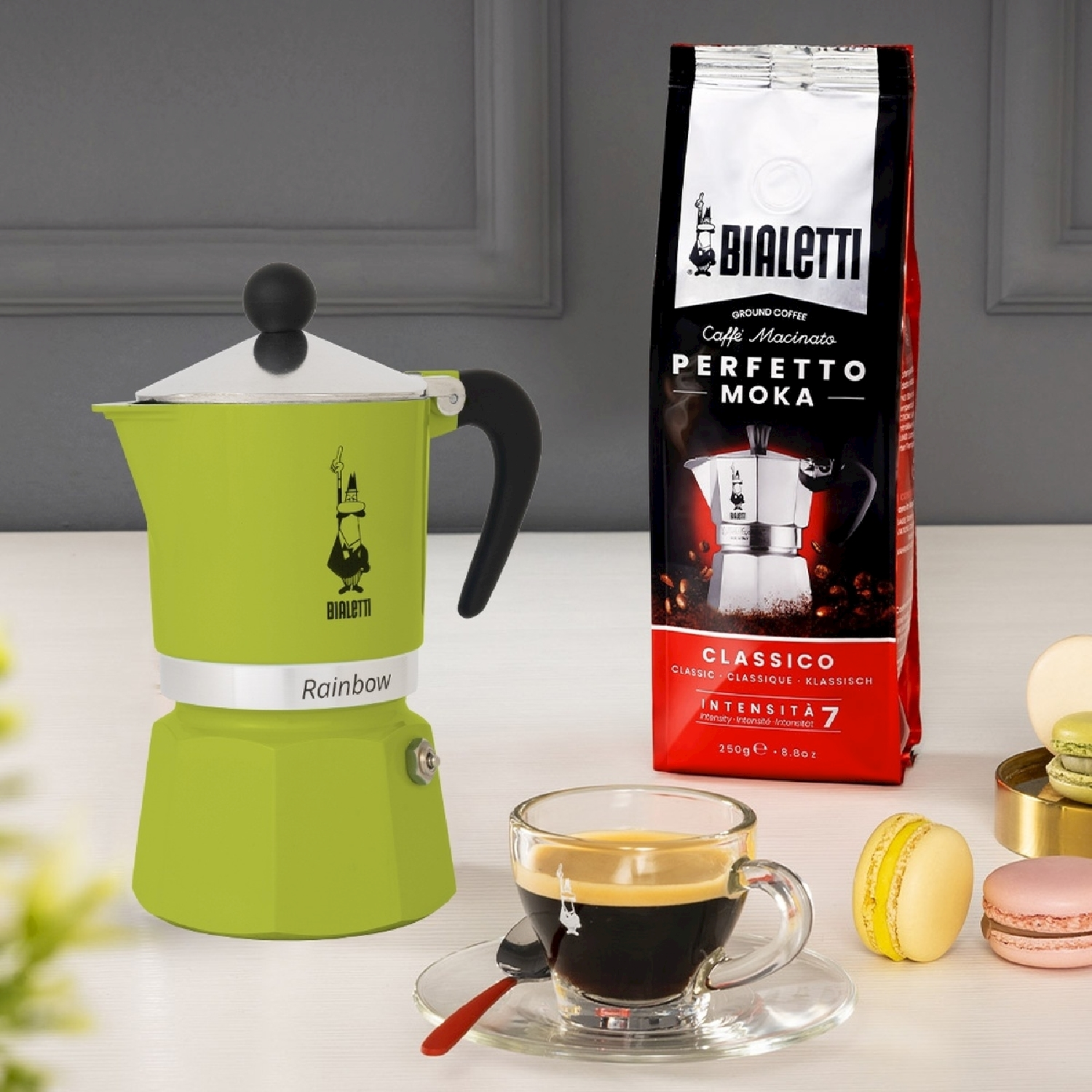 Una Bialetti Rainbow verde, una tazza di caffè, un sacchetto di caffè e macaron su una superficie bianca. Sfondo un muro e un fiore sfocato.
