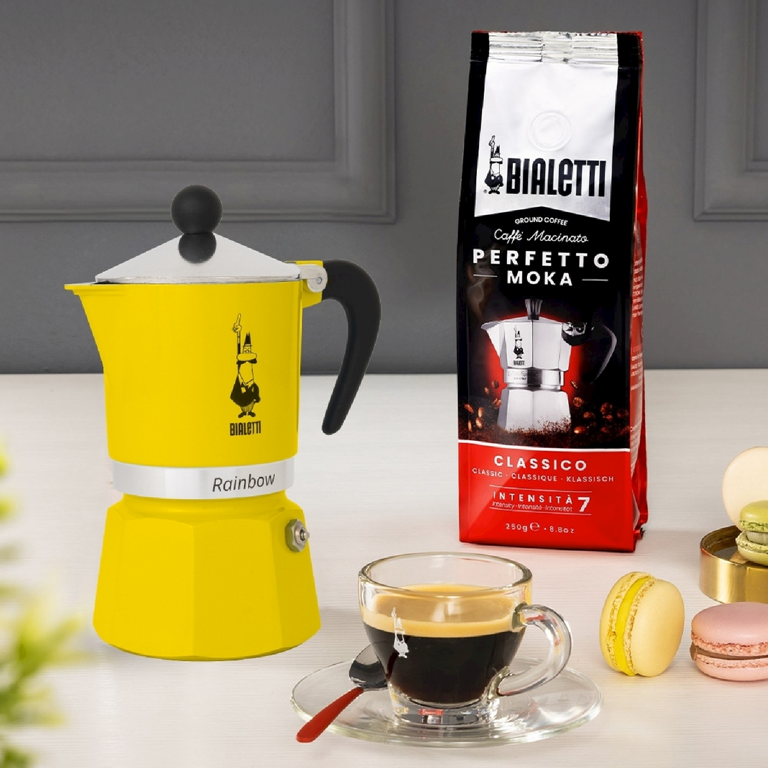 Una caffettiera moka Bialetti gialla, un sacchetto di caffè, una tazza di caffè e macarons sono su un tavolo. Lo sfondo è un muro.