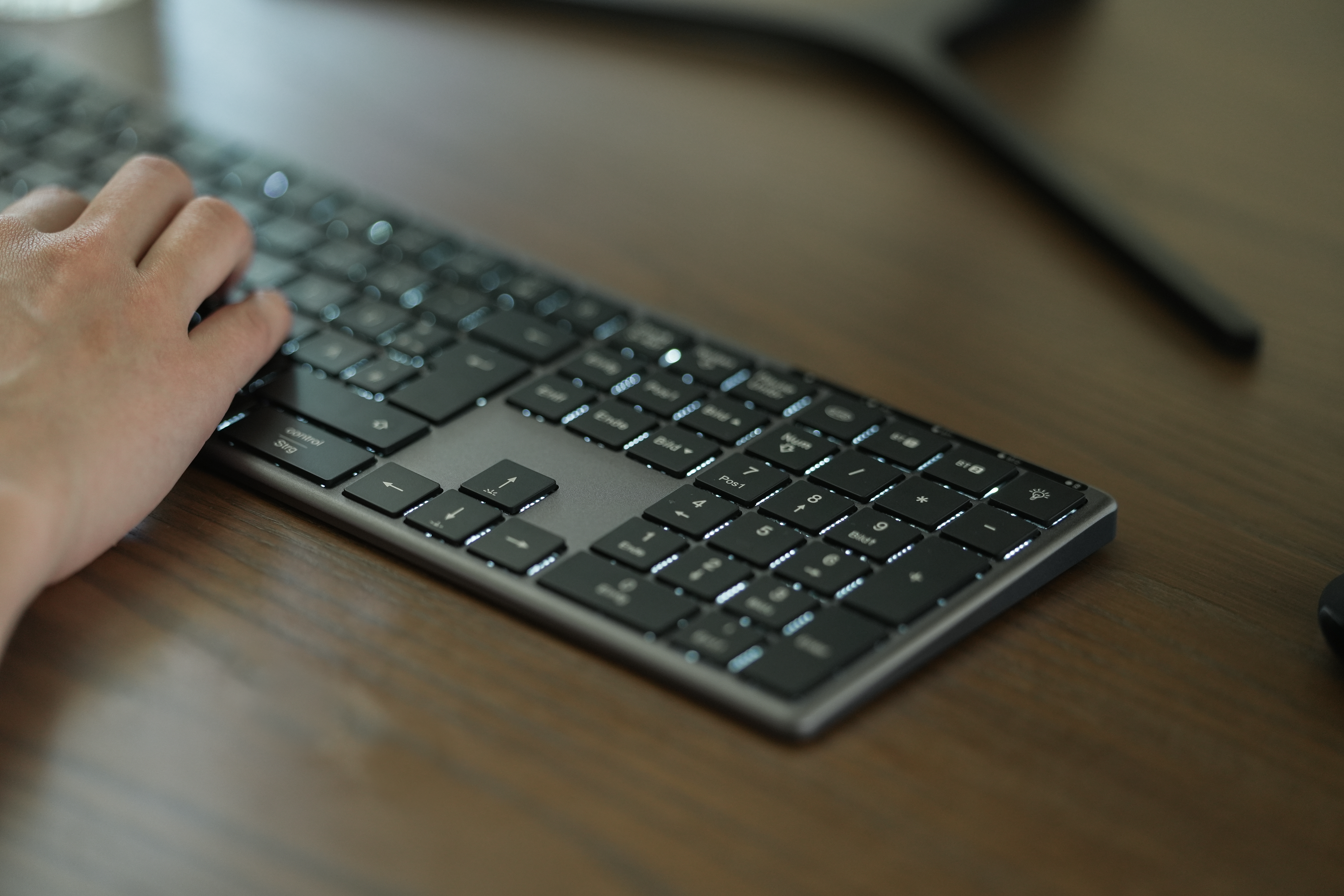 Eine Hand tippt auf einer schwarzen Tastatur auf einem Holzschreibtisch. Die Tastatur hat beleuchtete Tasten.