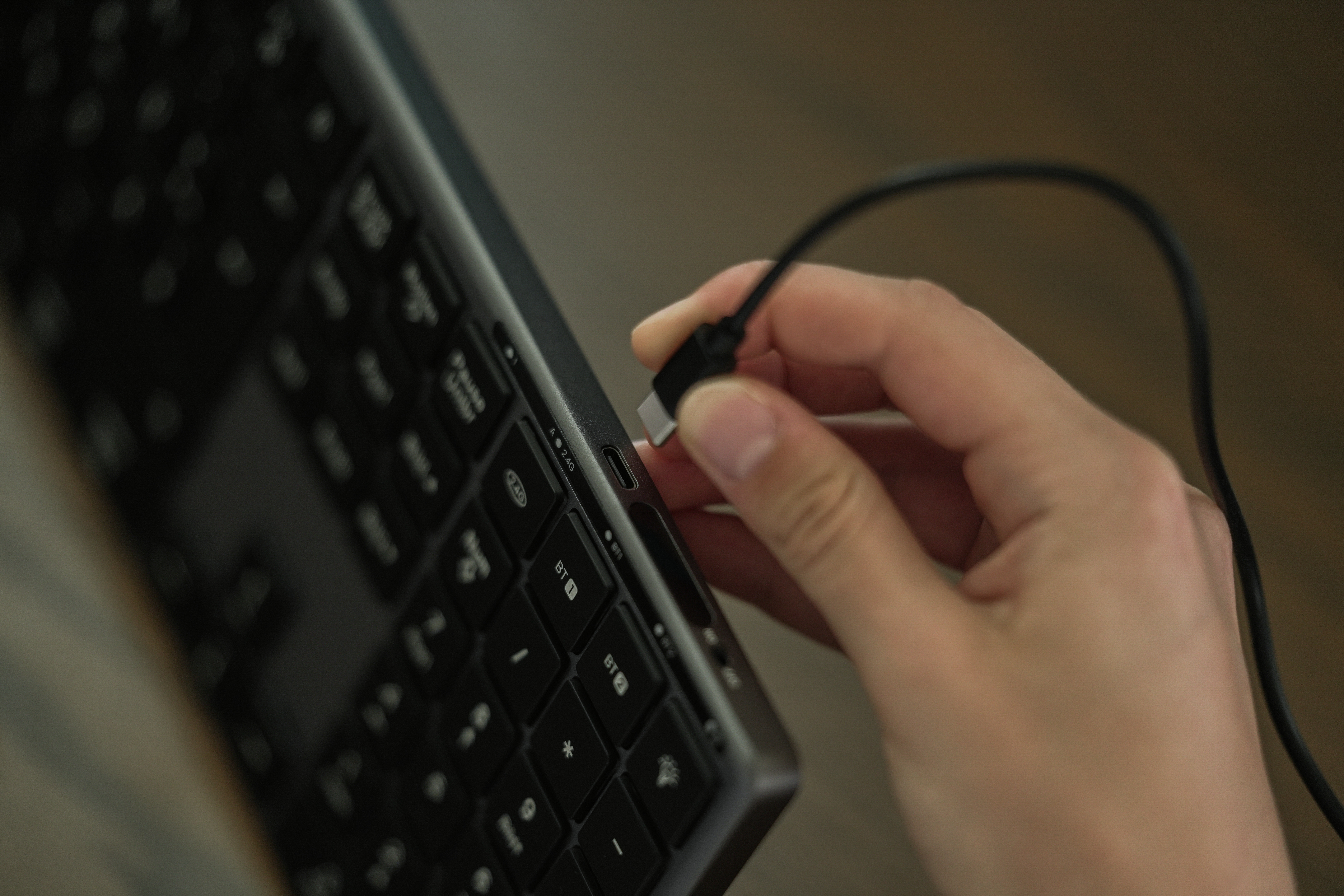 Eine Hand steckt ein USB-C-Kabel in eine Tastatur. Die Tastatur ist schwarz, das Kabel ist schwarz, der Hintergrund ist unscharf.
