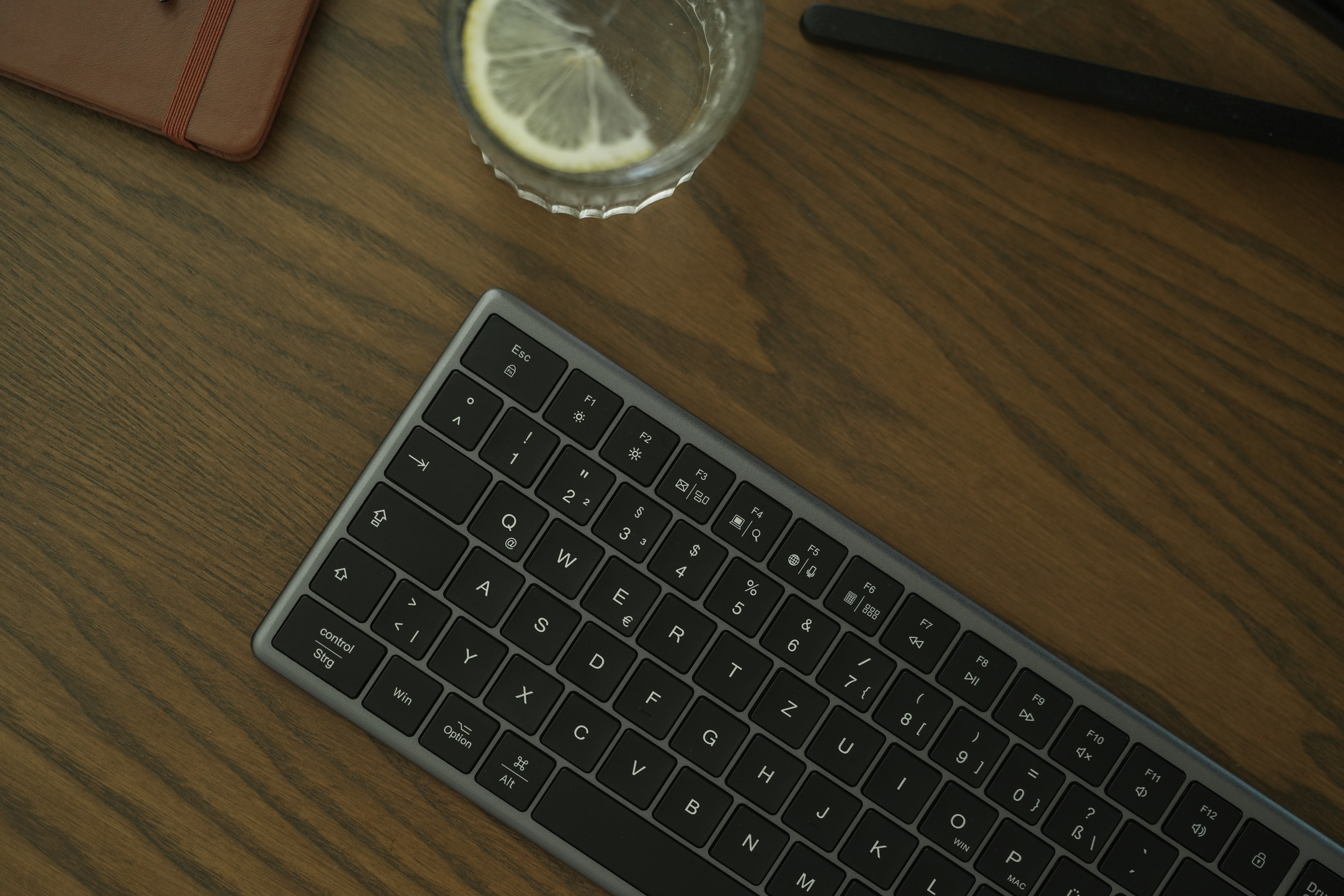 Tastatur, Glas Wasser mit Zitrone und Notizbuch auf Holzschreibtisch. Die Tastatur ist grau.
