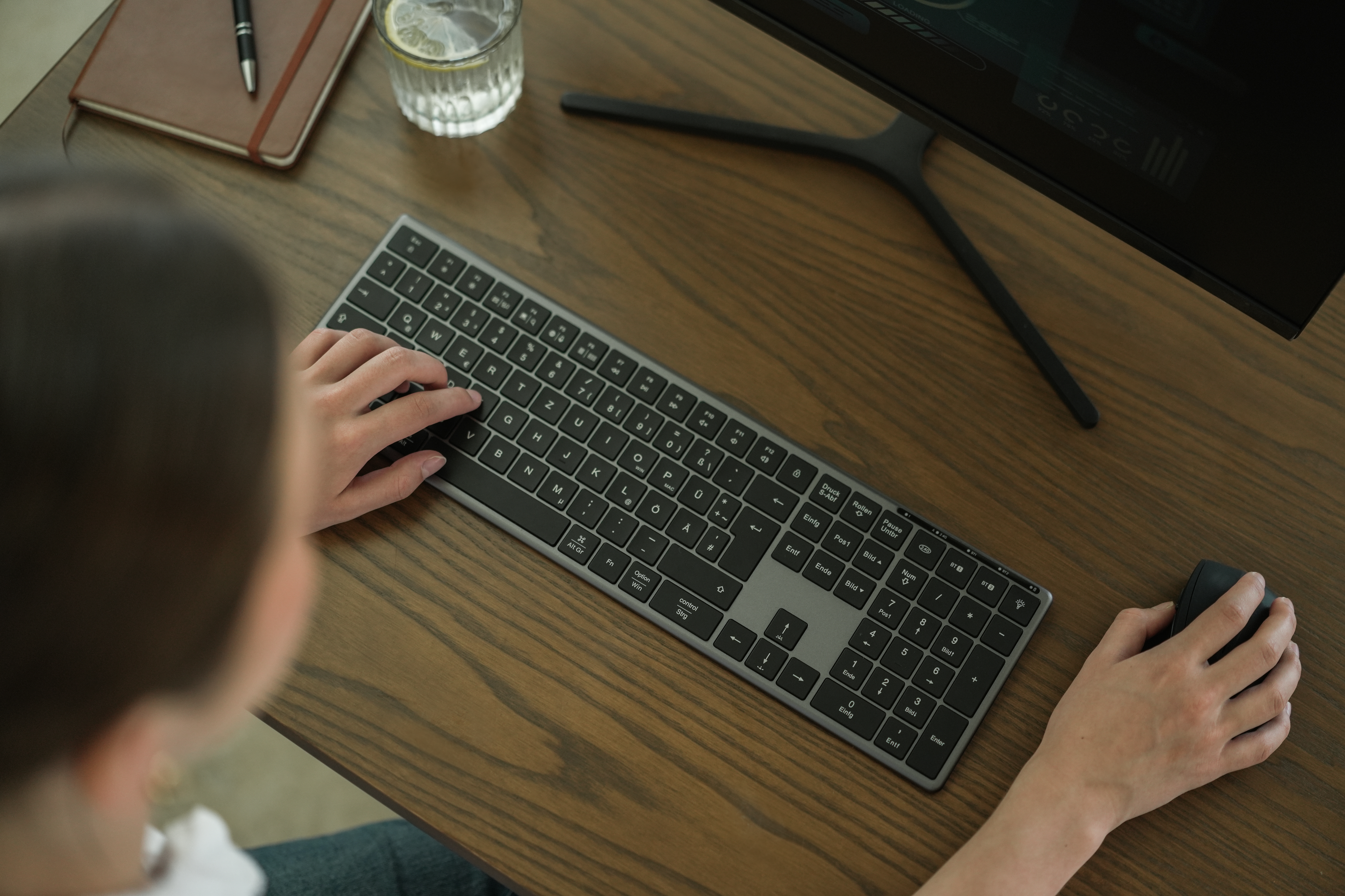 Person benutzt Tastatur und Maus auf einem Holzschreibtisch. Ein Monitor und andere Gegenstände sind ebenfalls vorhanden.