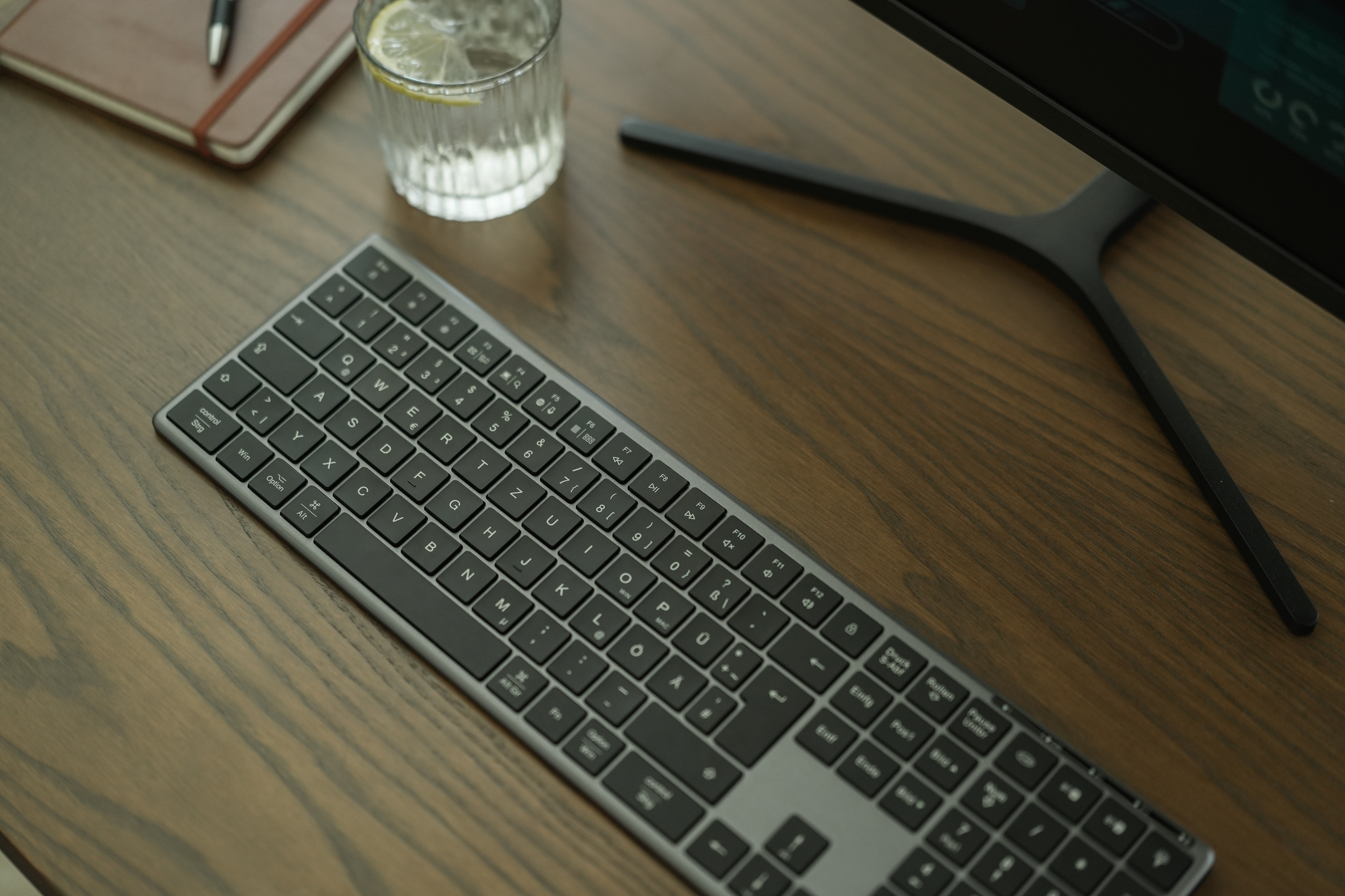 Tastatur, Glas Wasser mit Zitrone, Stift und Notizbuch auf Holzschreibtisch. Monitorständer sichtbar.
