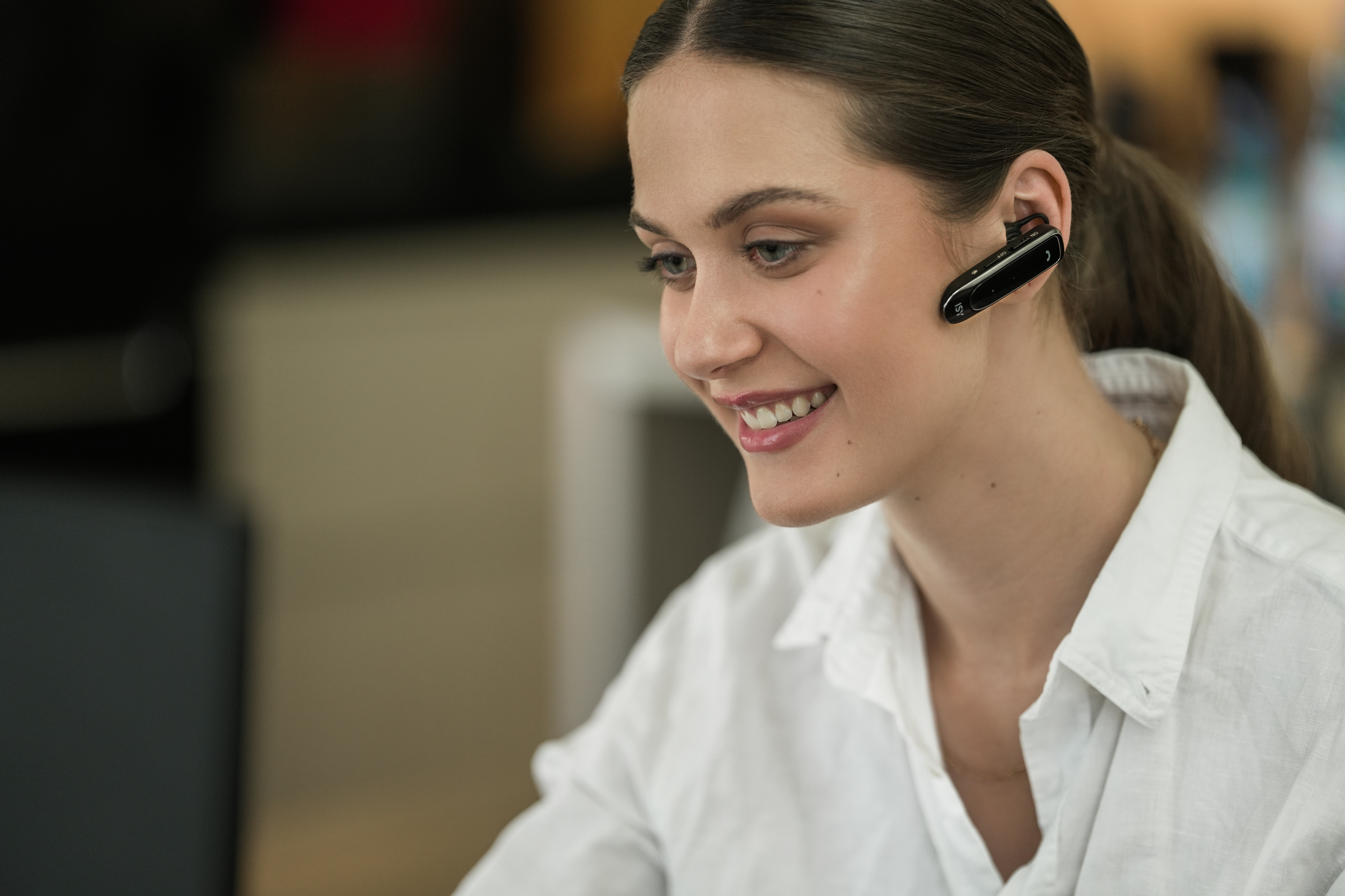 Eine lächelnde Frau mit Headset. Sie trägt ein weißes Hemd. Der Hintergrund ist unscharf, neutrale Farben.