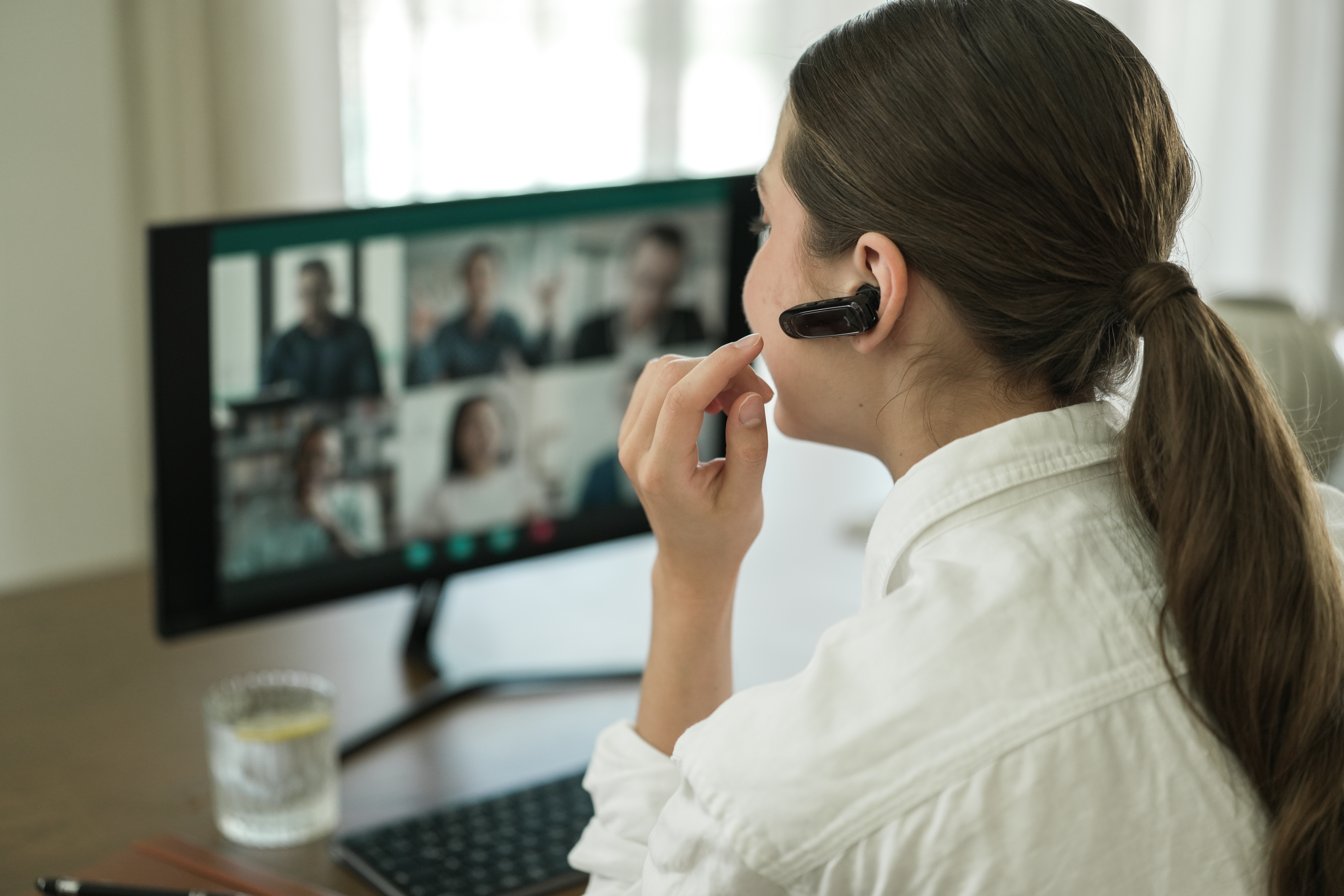 Frau im weißen Hemd bei einem Videoanruf mit Headset. Bildschirm zeigt weitere Teilnehmer.